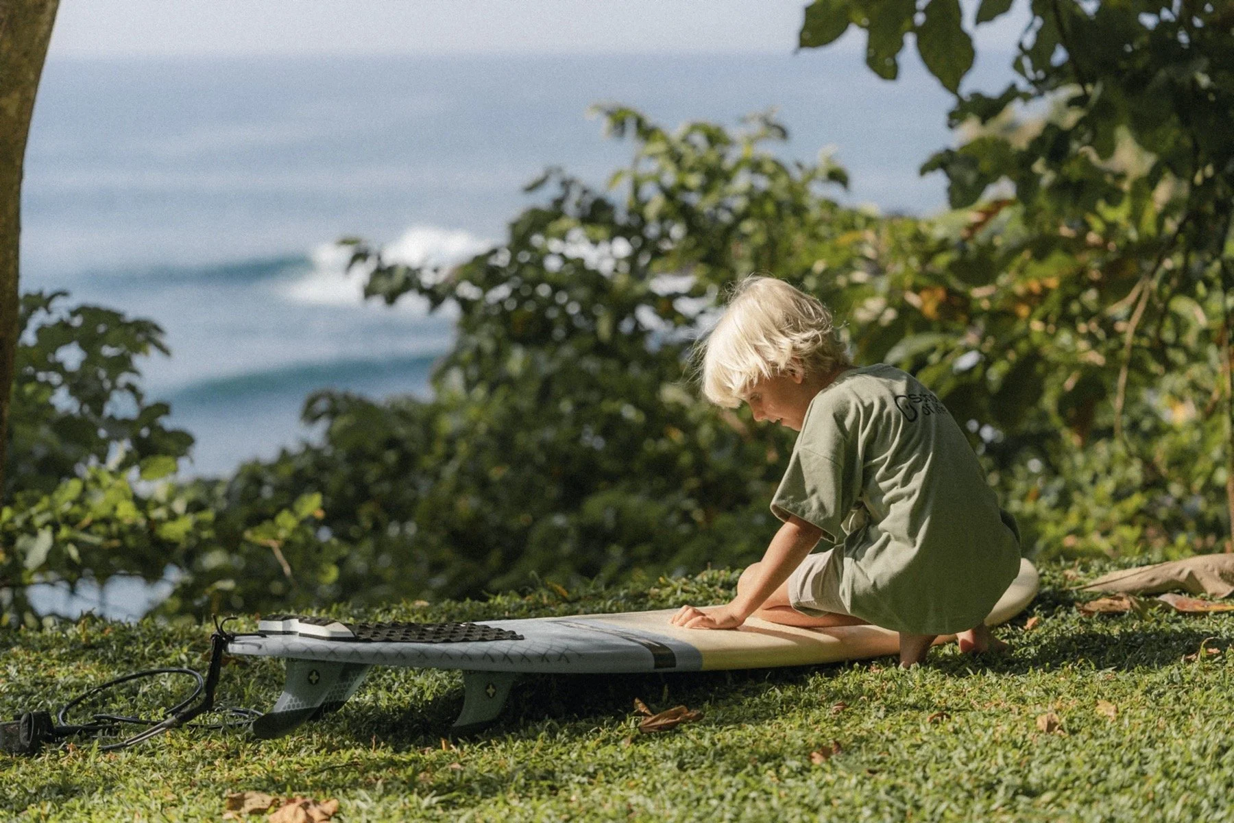 Child sitting on grass next to a surfboard near the ocean