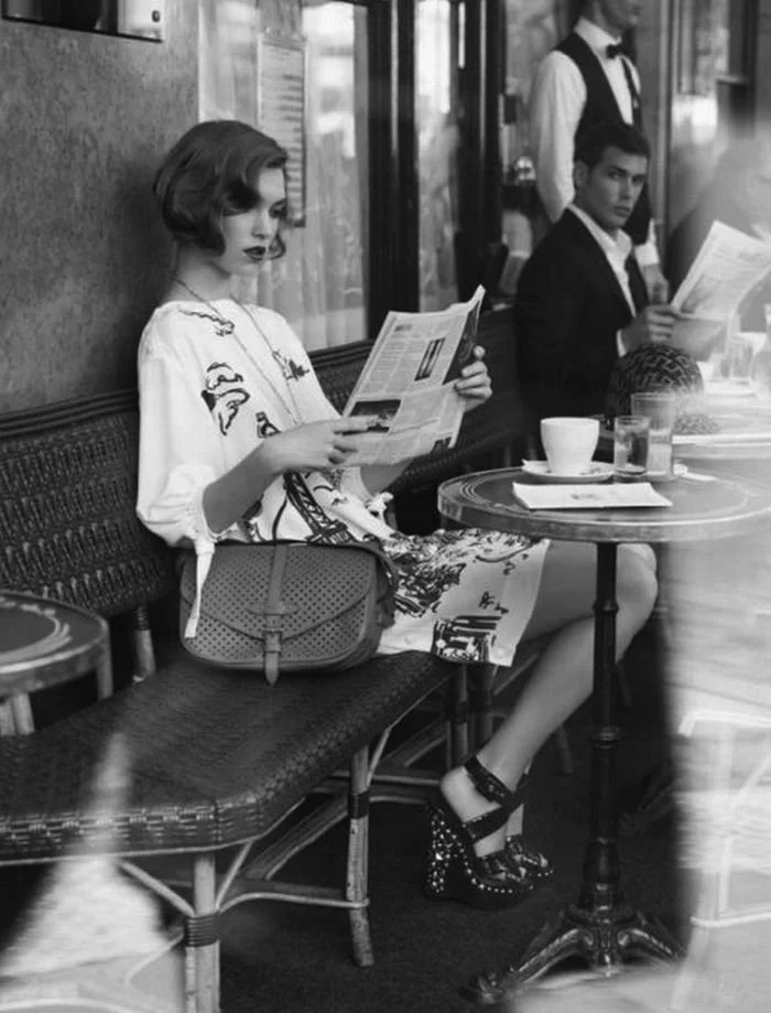 A woman with short hair and a patterned dress sitting in a cafe, reading a newspaper, with a purse on her lap, and cups on the table. A man in a suit is sitting at the next table looking at her. A waiter stands in the background.