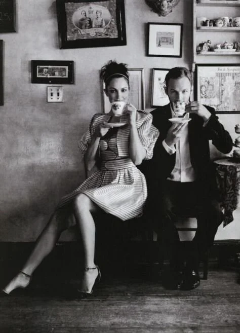 A man and woman sitting on a bench in a vintage-style room, drinking from cups, with framed pictures on the wall behind them.