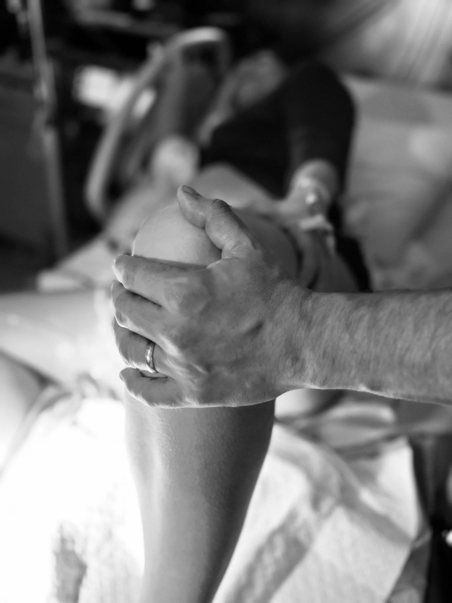 A black and white close up photo of a supportive partner's hand helping support and hold up his birthing partner's knee and leg during pushing phase of childbirth.