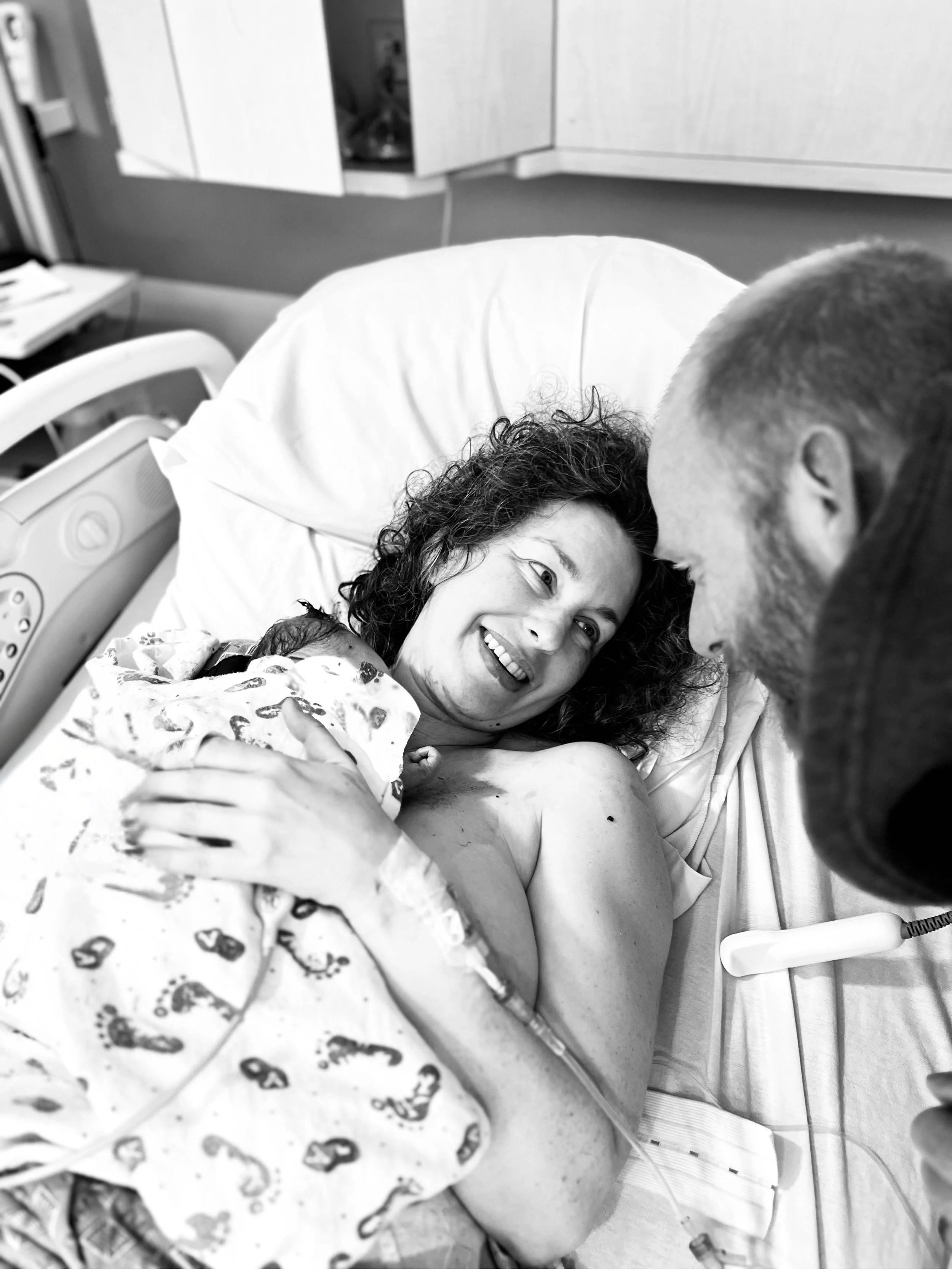A woman who just gave birth for the first time is holding her newborn baby and smiling at her husband who is leaning over her, in a hospital room.