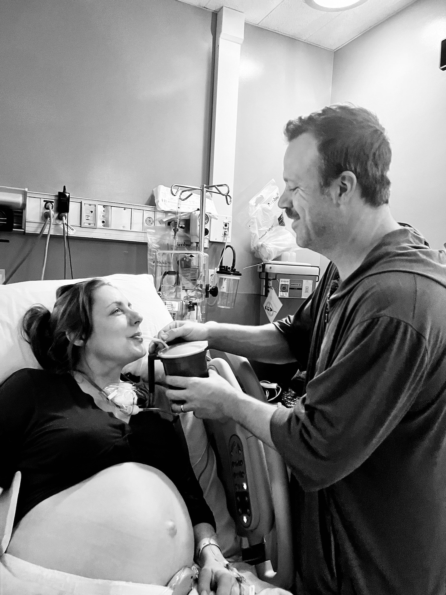 The parter of a laboring woman offering her sips of water from a straw while she rests in a hospital bed smiling and looking back at her partner beside her. The man is feeding her with a straw, smiling back.