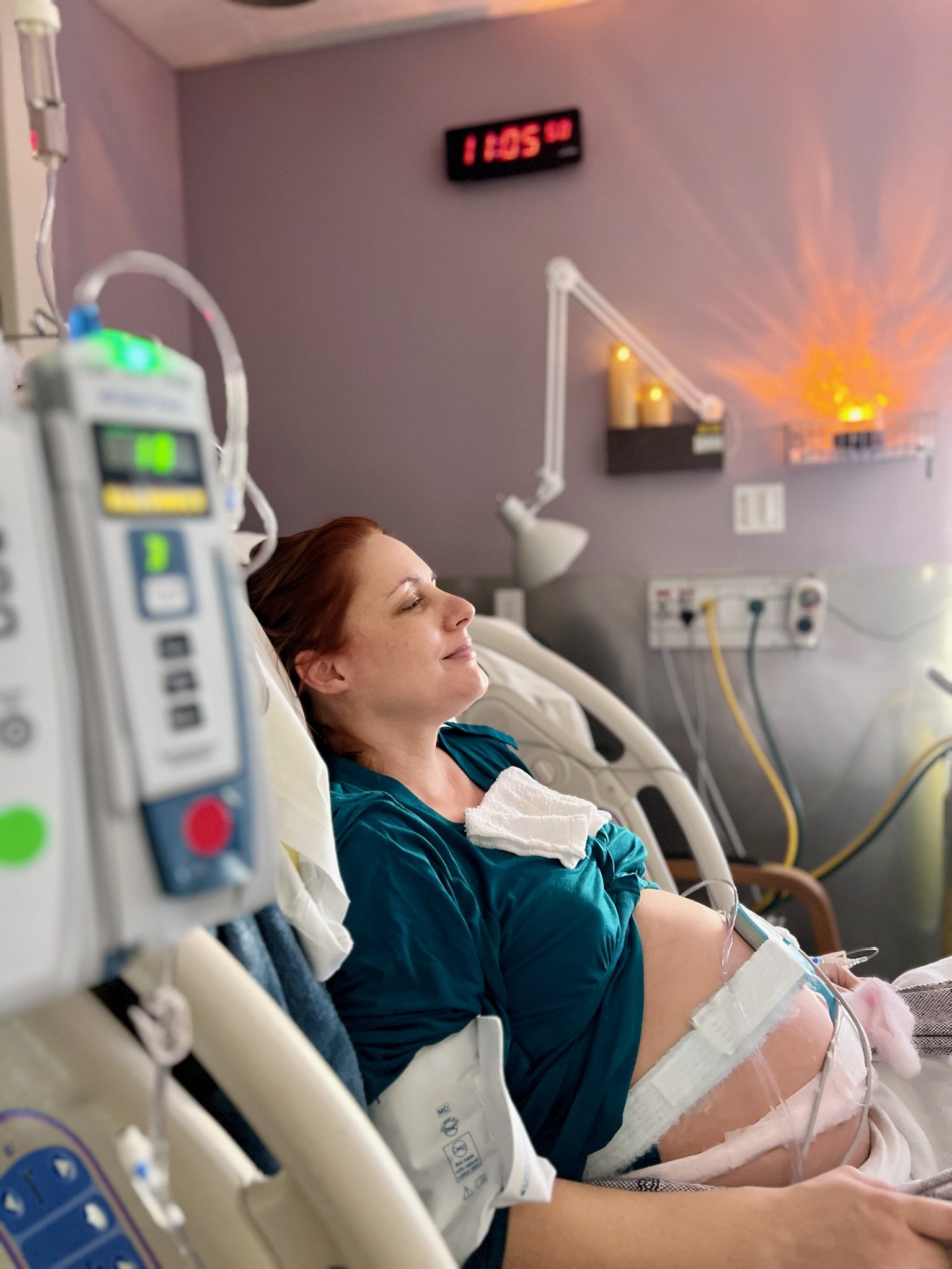 A woman in labor is resting upright in a hospital bed, smiling with her eyes closed, with medical equipment around her, in a hospital room with a digital clock showing 11:05 and candles on the wall.