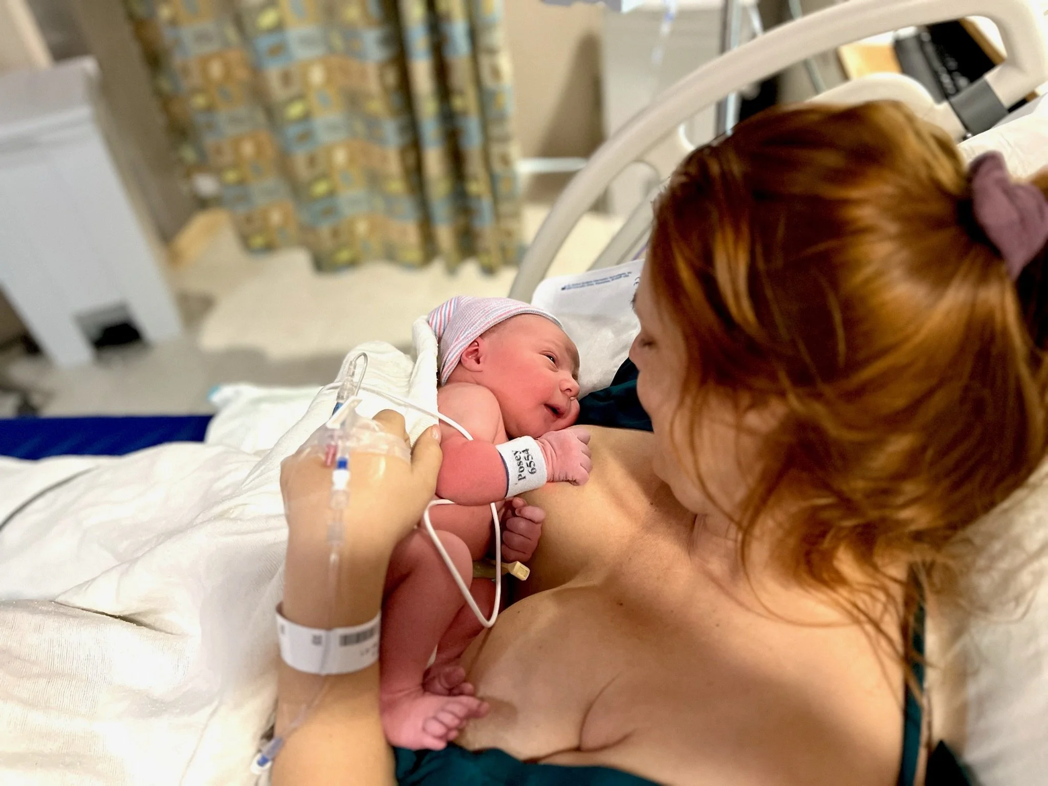 Newborn baby lying on mother's chest in hospital bed, with medical tubes attached, both looking at each other.