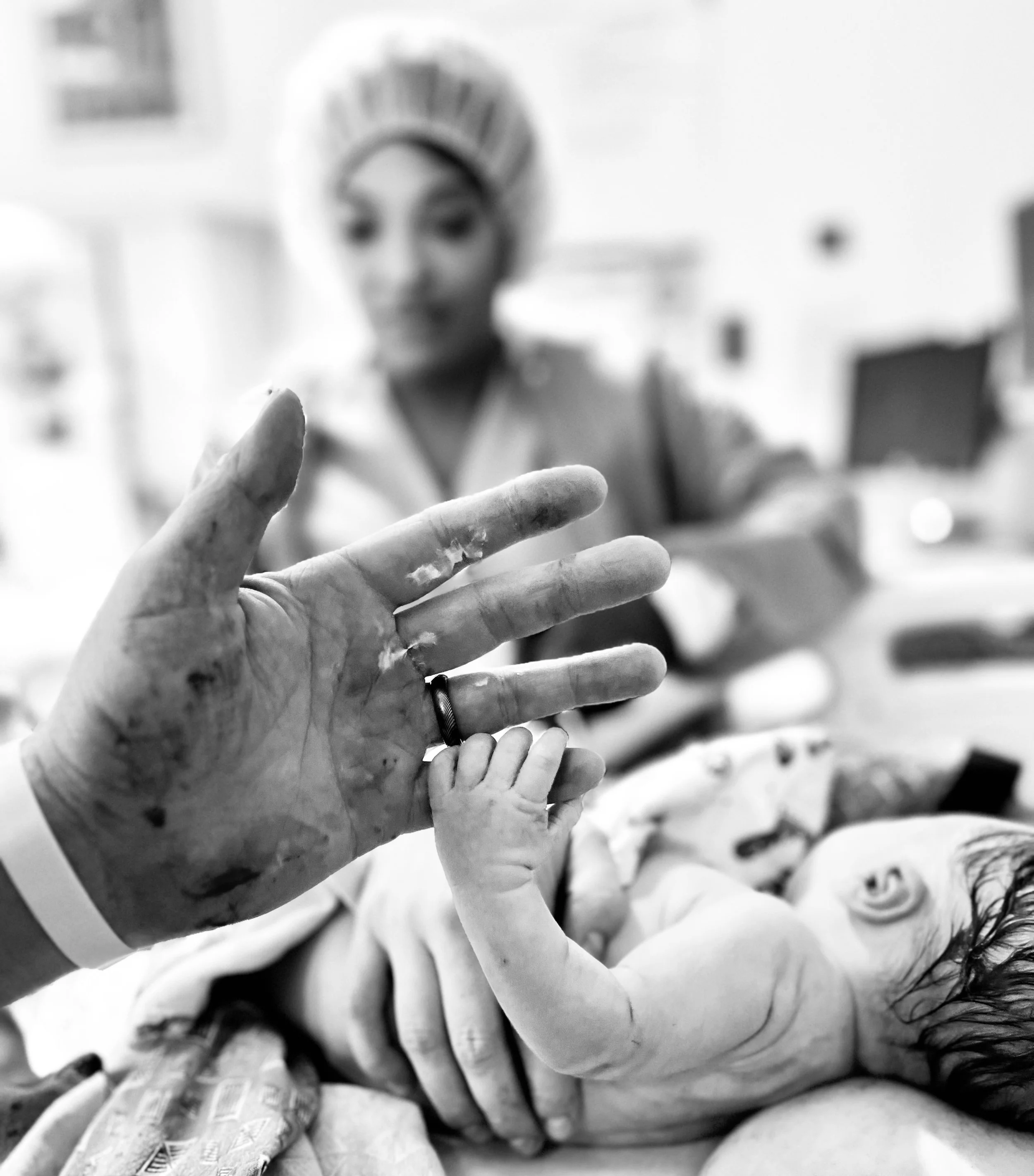 Black and white photo of a freshly born baby's hand grabbing the finger of her dad. The dad's hands have blood and vernix on them from helping bring baby up to mom's chest