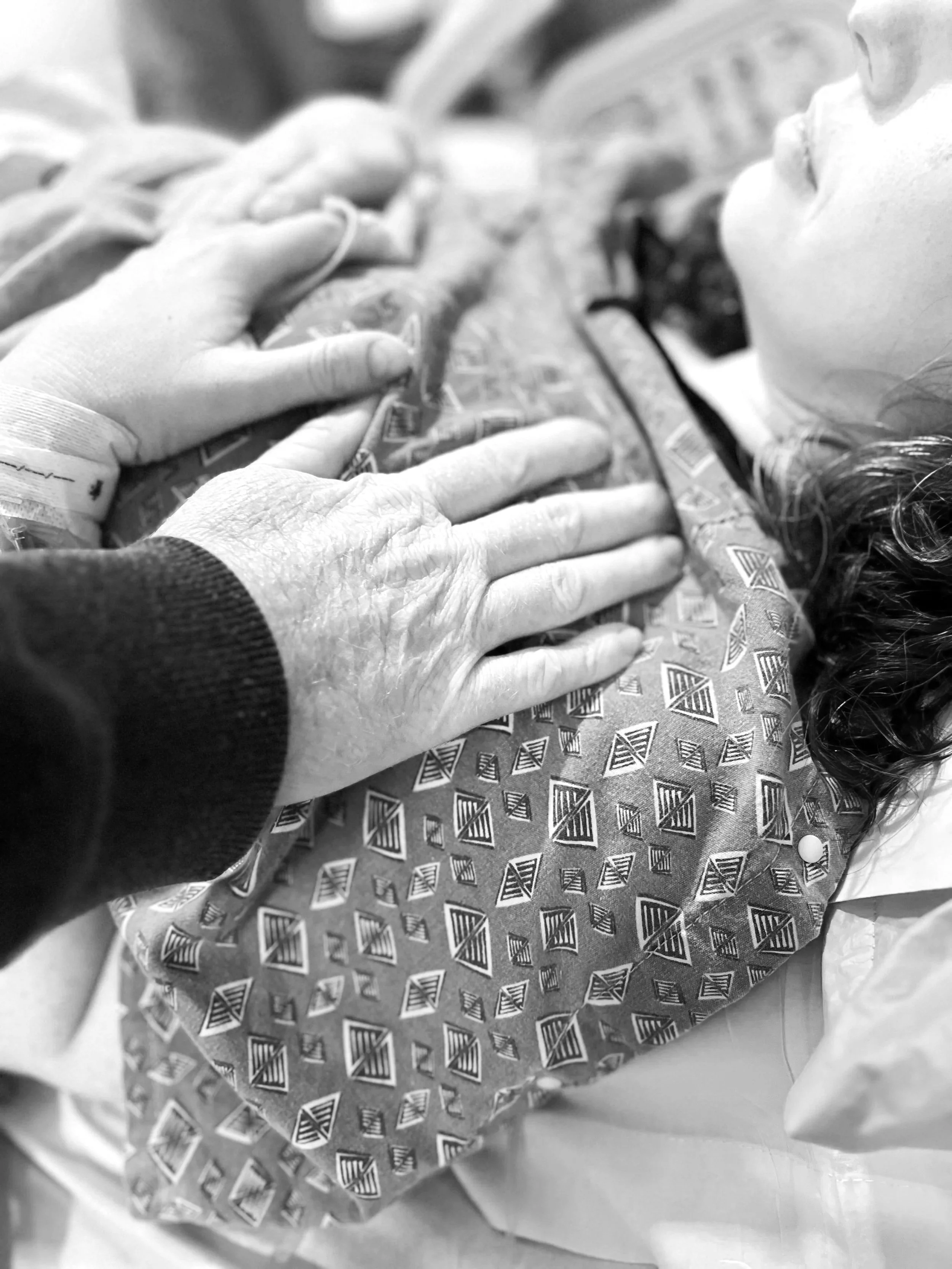 A laboring woman is lying in a hospital bed with her husband gently placing a hand on her chest, indicating a moment of comfort or care.