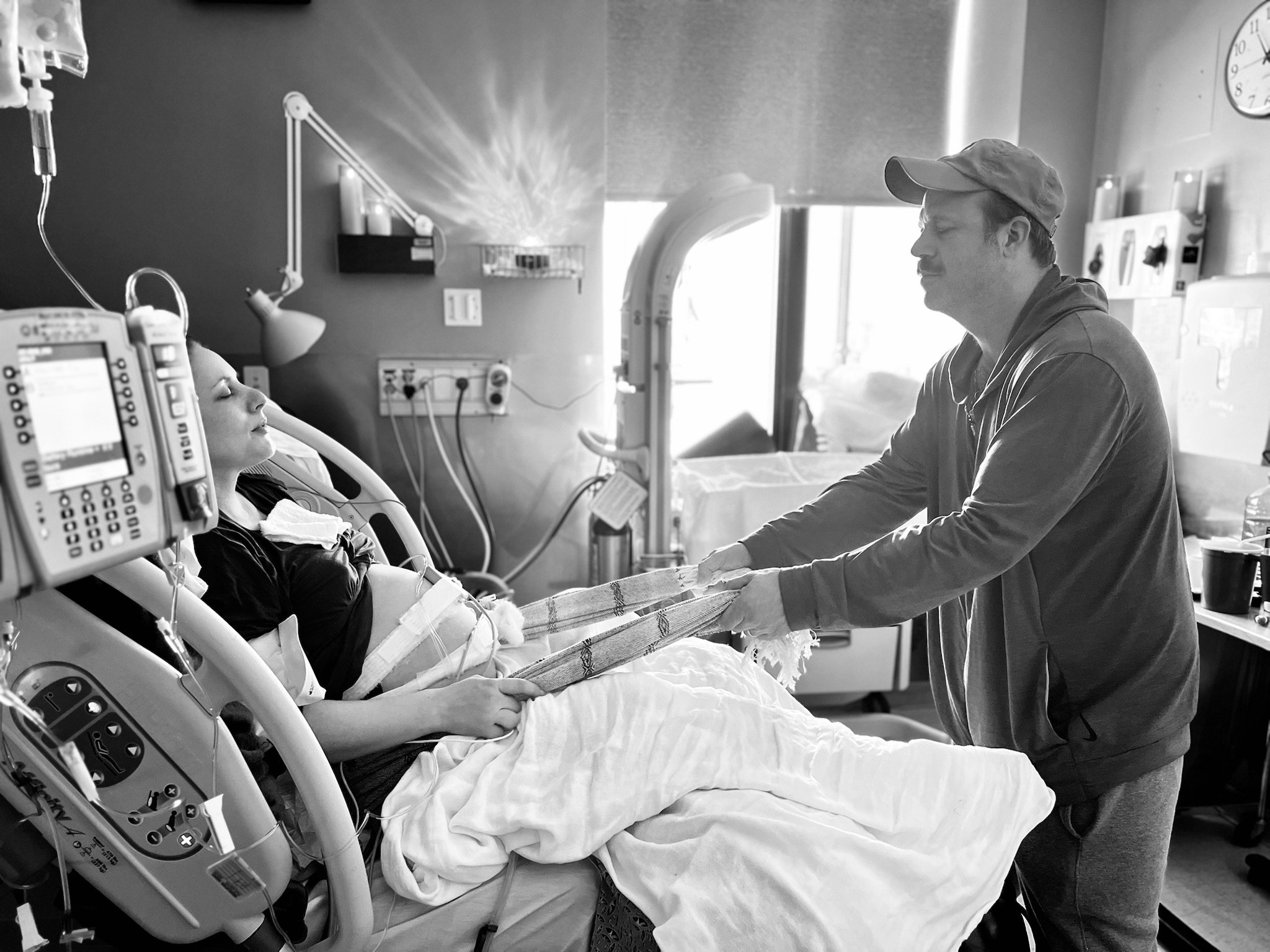 A laboring woman in a hospital bed being supported by her husband using the Rebozo in a gentle sifting motion. The scene is in black and white.