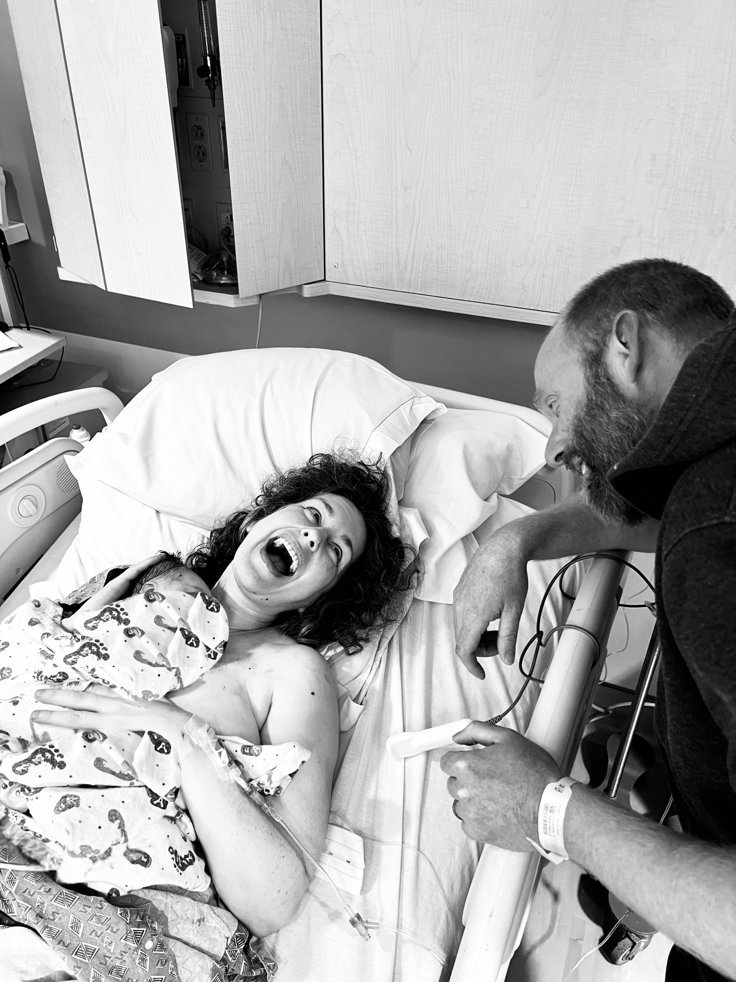 A woman who just gave birth is holding her brand new baby skin to skin on her chest, smiling and laughing, with her husband smiling back gazing at their newborn baby. 