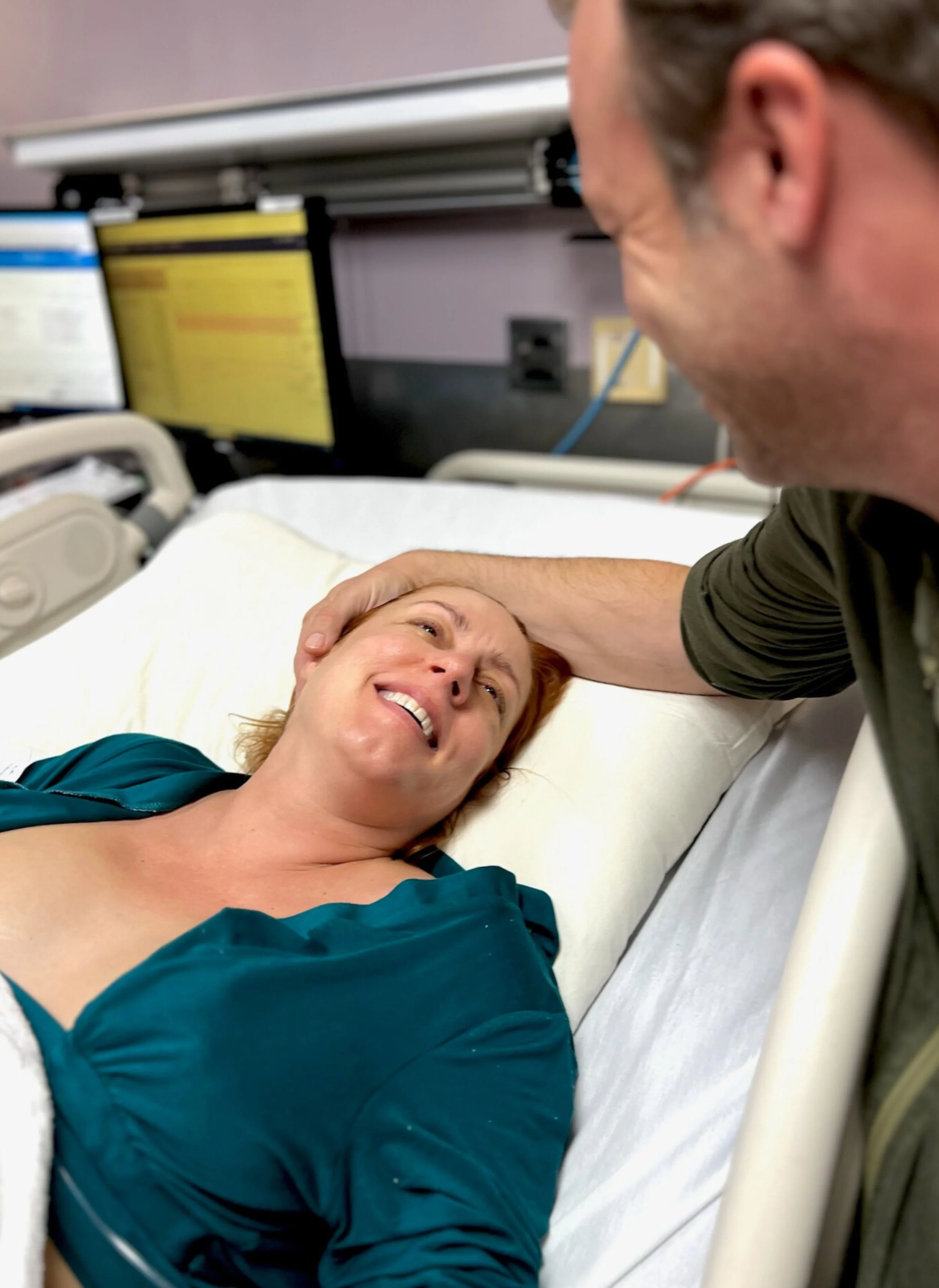 A laboring woman lying in a hospital bed smiling and looking her husband who is gently touching her forehead. The woman is about to meet her baby and is happy.