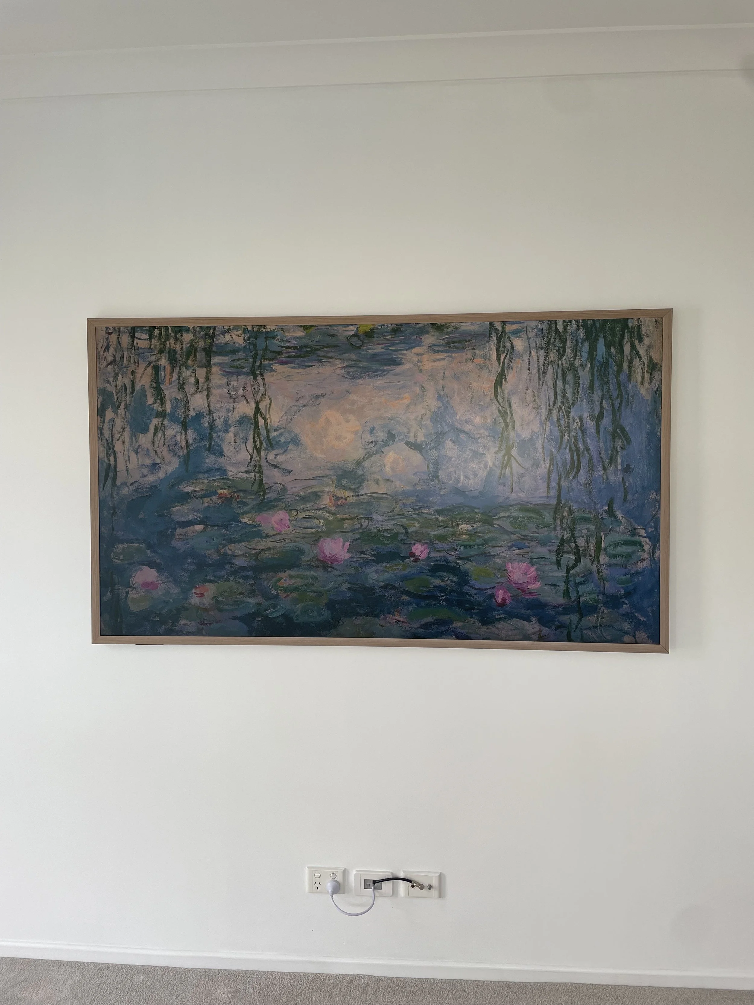 A framed painting of water lilies hanging on a white wall. Below the painting, there is a power outlet with a black power strip plugged into it.