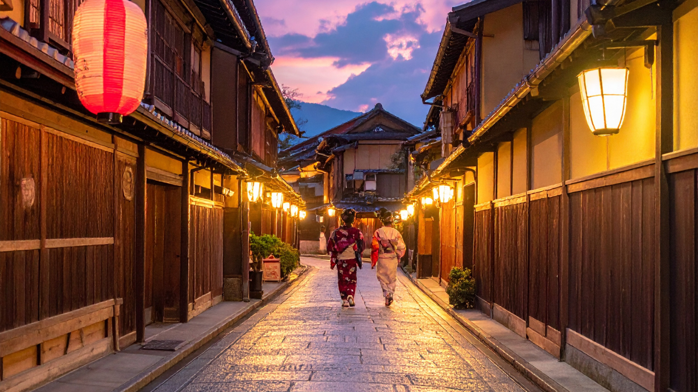 Two women wearing traditional kimonos walking down a lit alleyway with wooden buildings on each side at dusk.