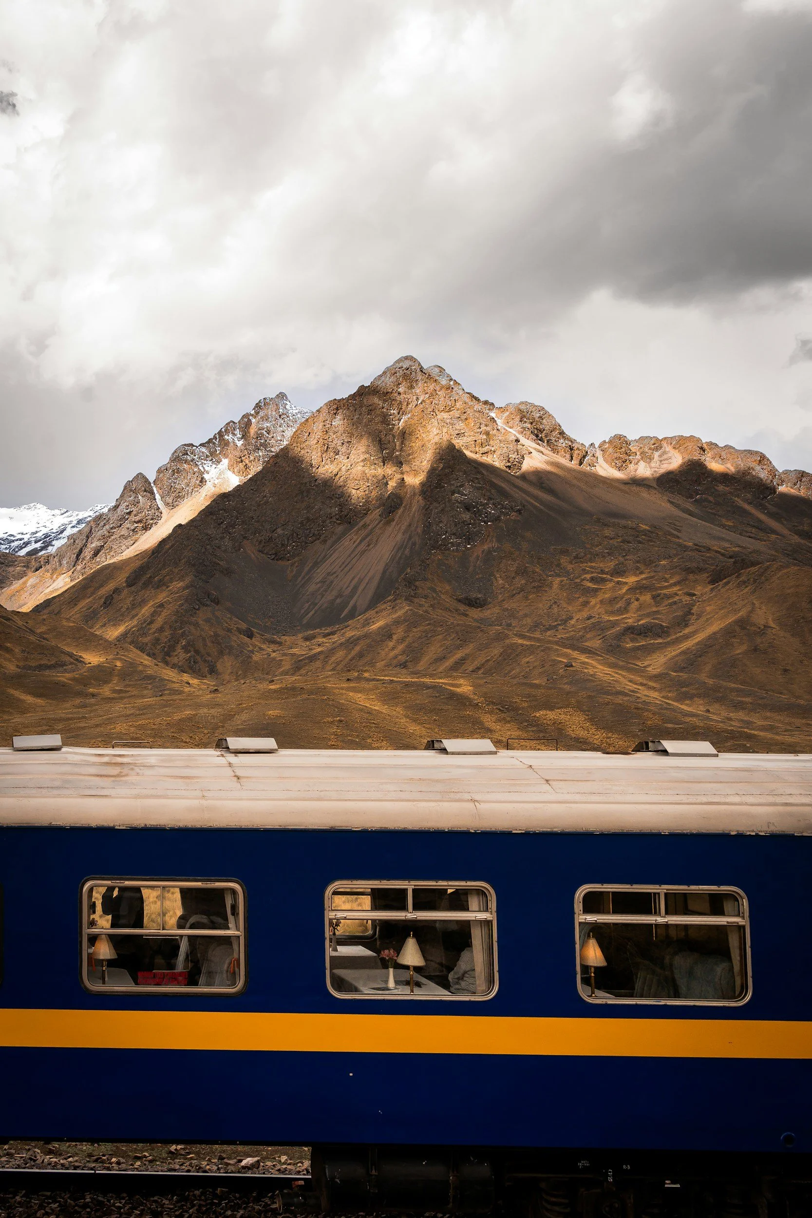 A blue and yellow train with three windows, with a mountain range in the background and cloudy sky overhead.