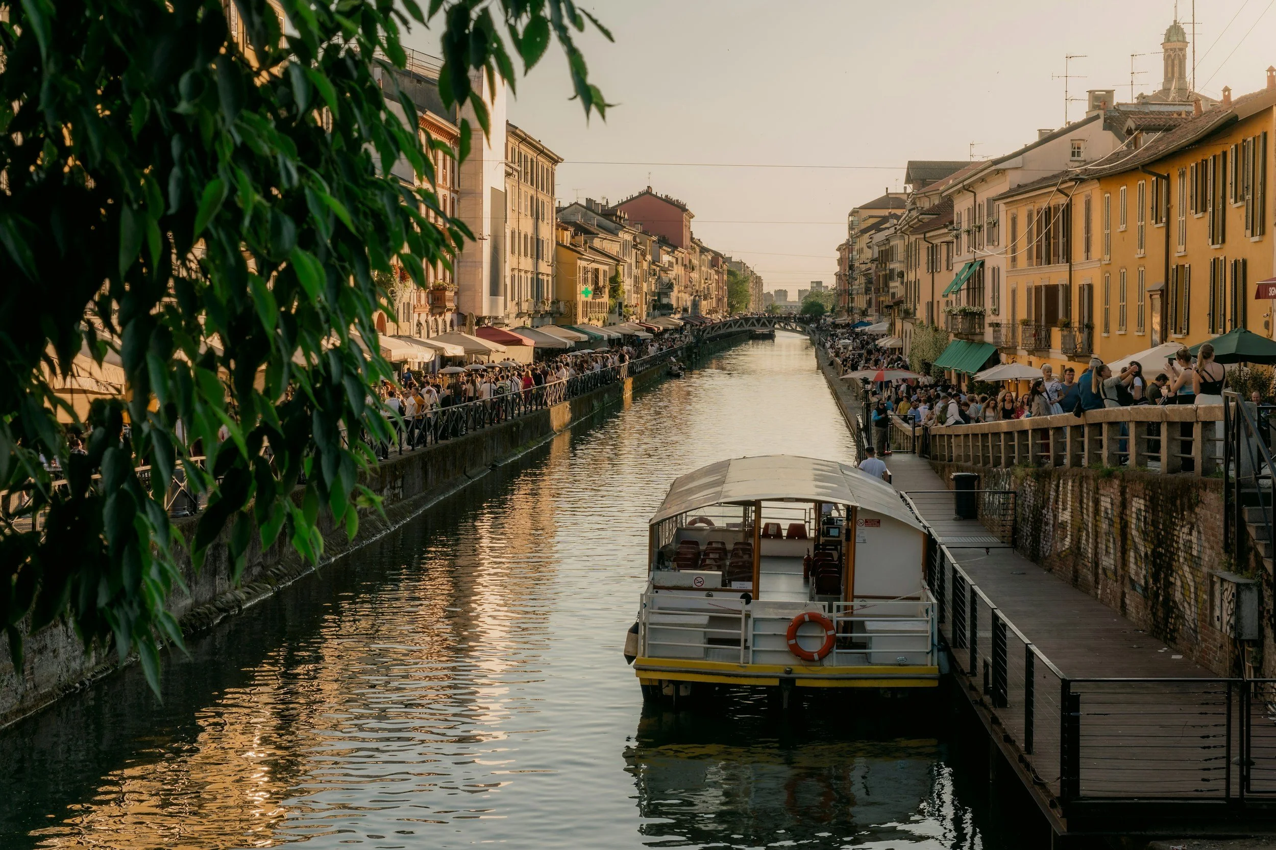 A canal lined with historic buildings and outdoor cafes, with a crowd of people walking along the water's edge in the evening light.
