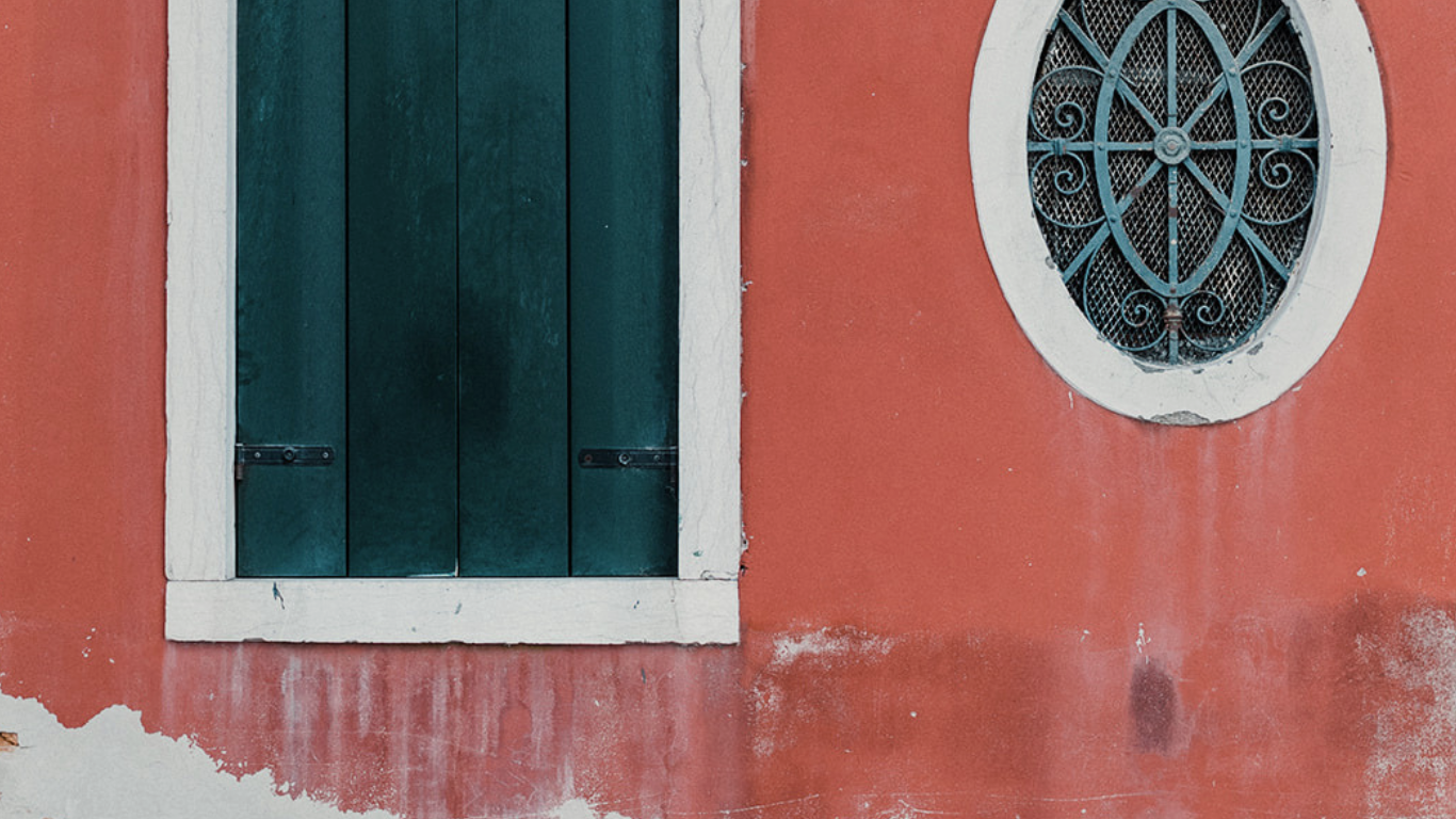 Terracotta red facade with teal shuttered window and oval ironwork detail, peeling plaster at the base.