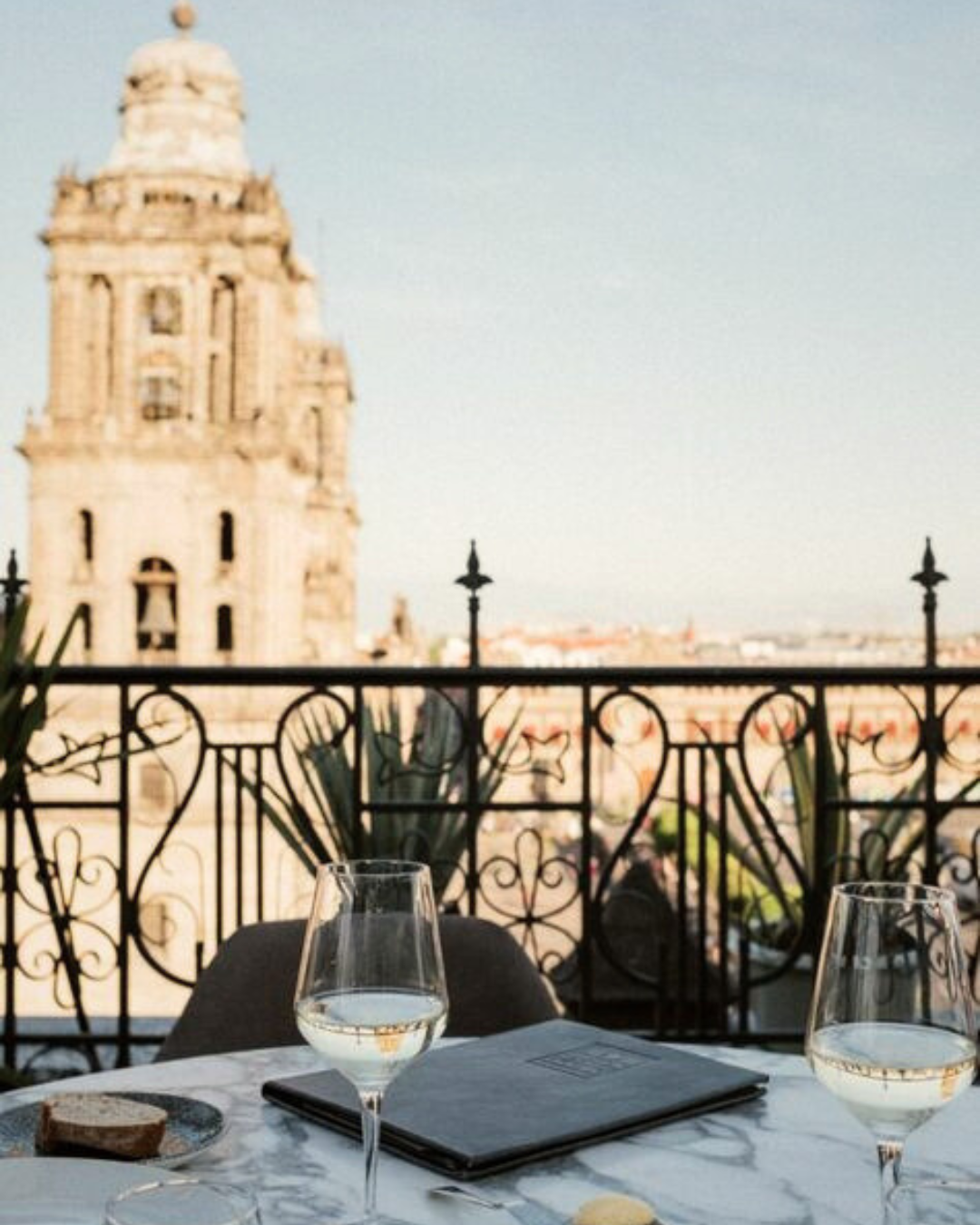A balcony table with two glasses of water, a menu, and a plate with bread. The background features an ornate wrought iron railing and a view of historic buildings and a church tower in a European city.