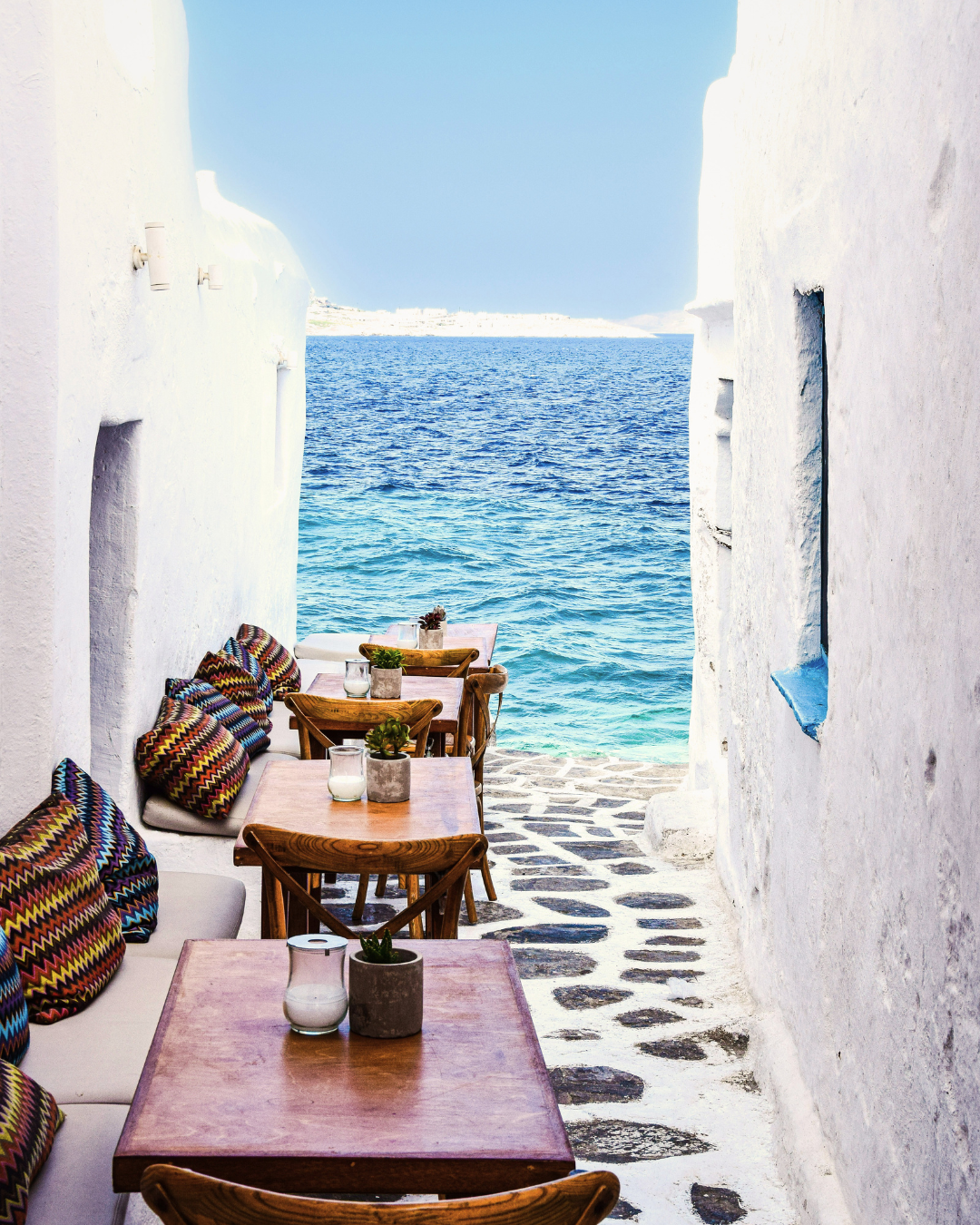 Outdoor seating area with wooden tables and colorful cushions on a white-washed wall, overlooking a blue sea under a clear sky.