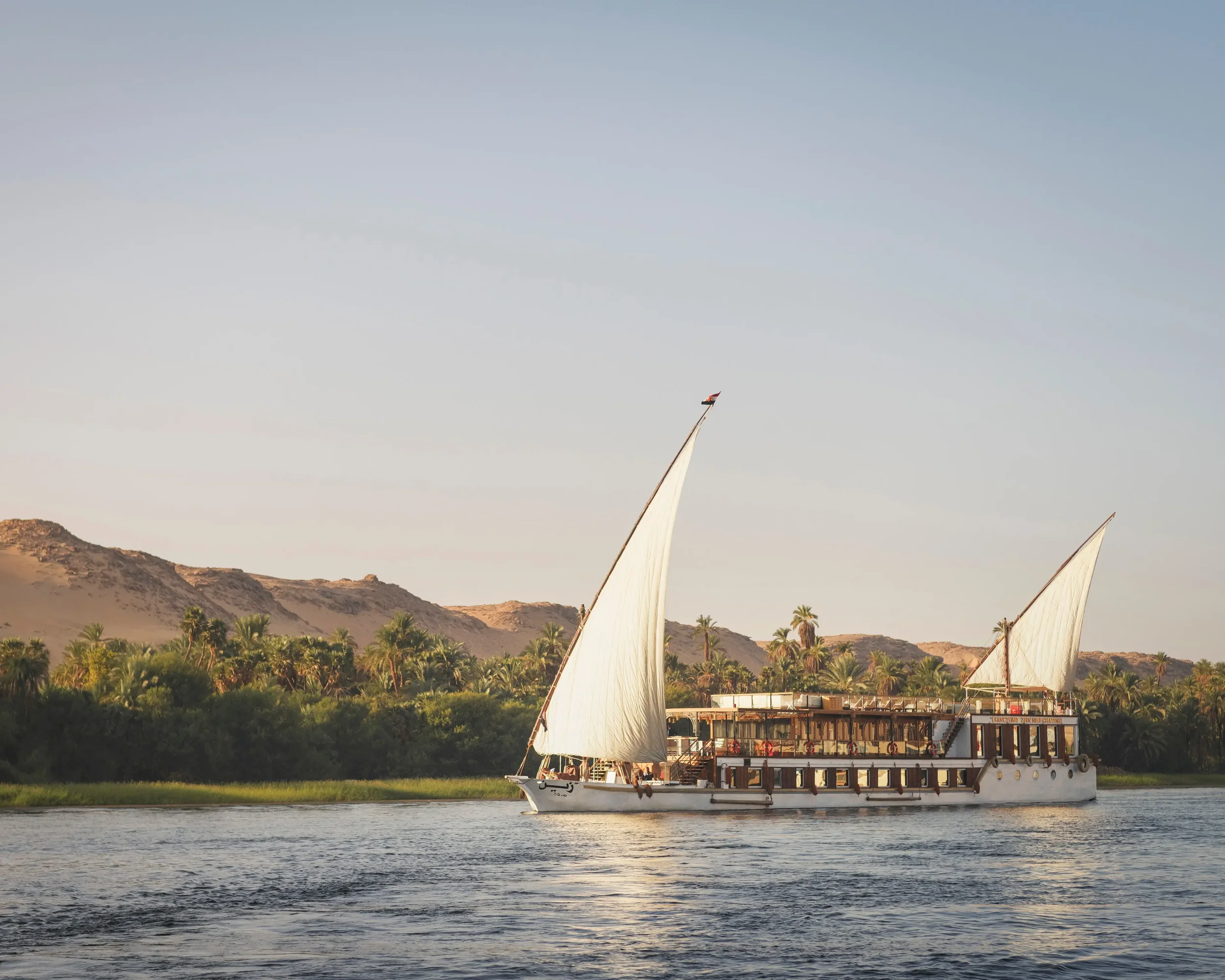 A traditional wooden sailboat with white sails sailing on the Nile river, with lush green trees and desert hills in the background under a clear sky.