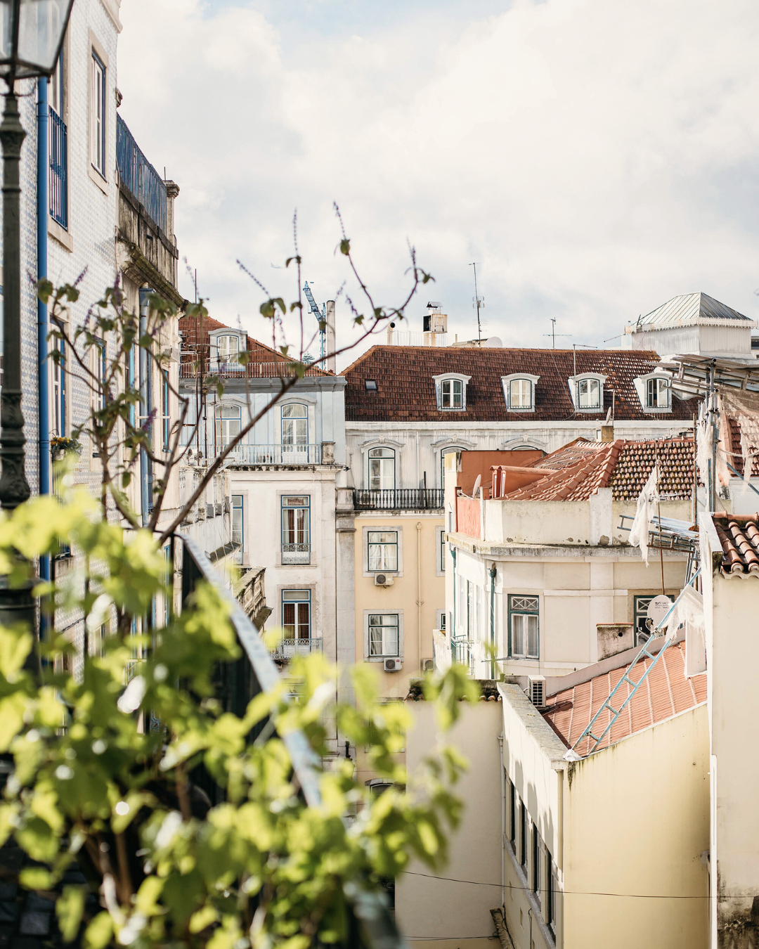 View of several old European-style buildings with white and pastel-colored facades, balconies, and red-tiled roofs, with some plants and laundry hanging outside on a balcony in the foreground under a partly cloudy sky.