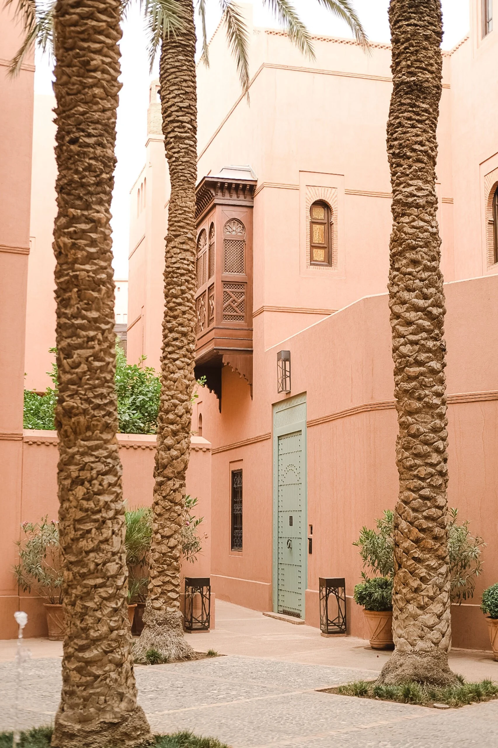 A courtyard with pink walls, green door, several palm trees, potted plants, and lantern-style light fixtures.
