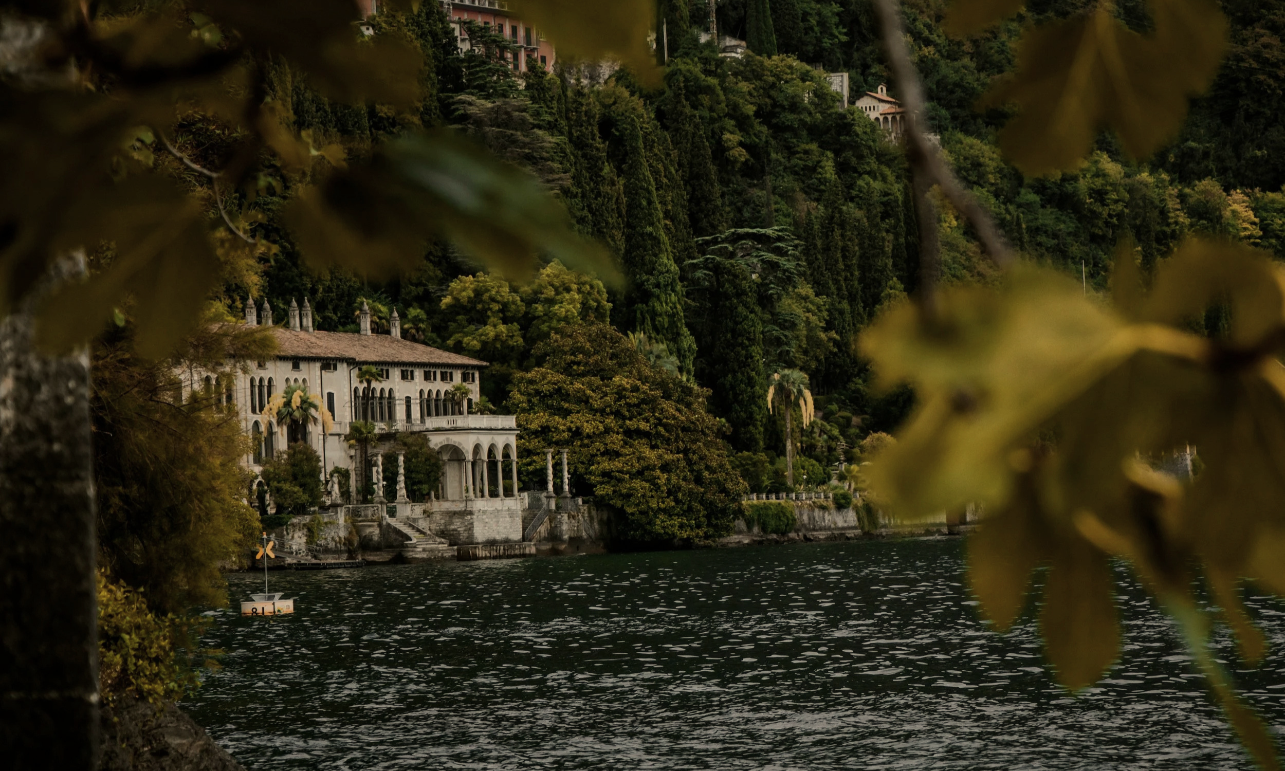 A lakeside view of a large, ornate white house on a hillside covered with dense green trees, with some buildings visible higher up on the hill. The house has arched windows and a tile roof, partially obscured by trees and leaves in the foreground. The water in the lake reflects the greenery and the house.