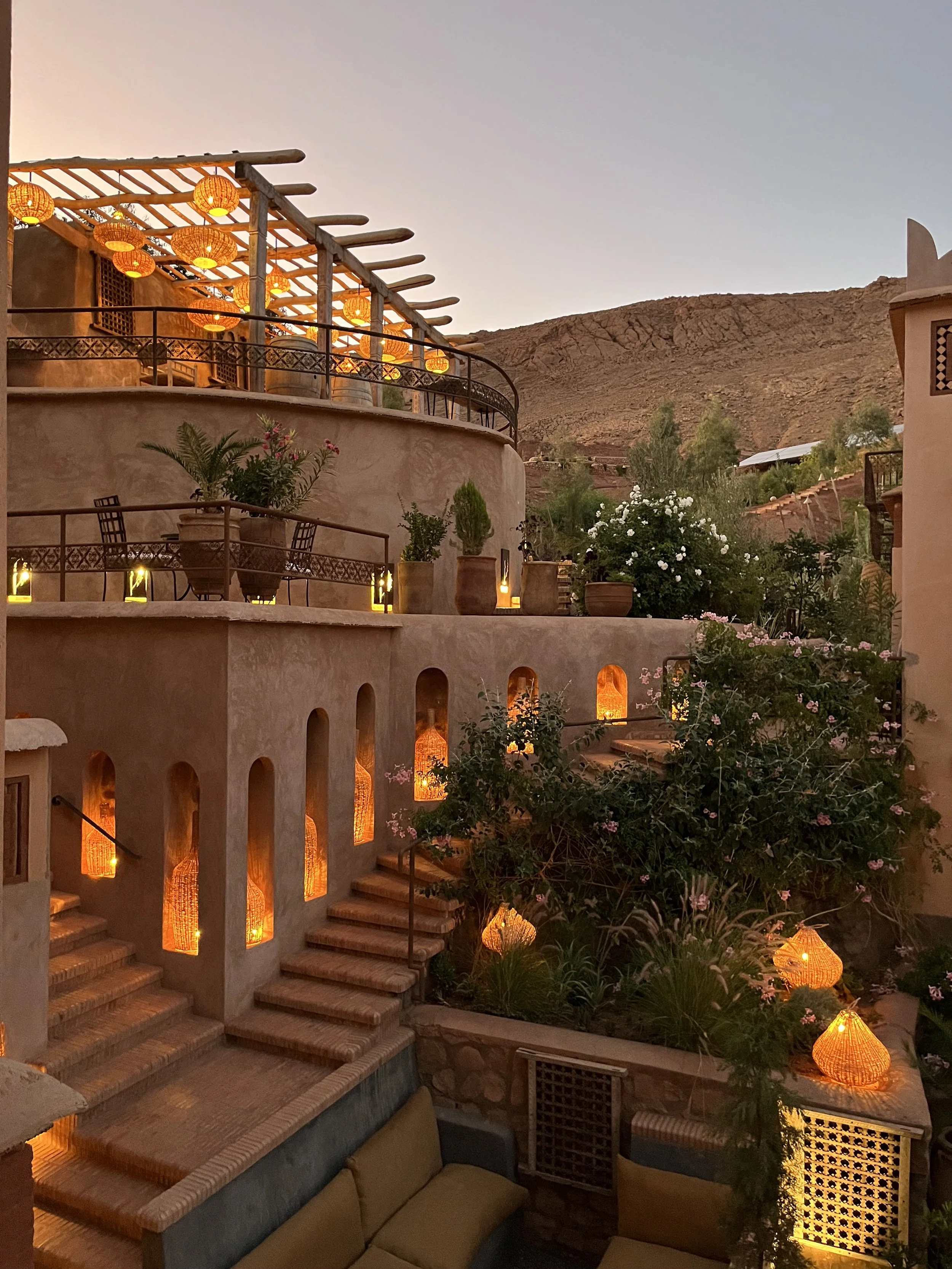 A multi-level outdoor terrace with terracotta walls, stairs, potted plants, and warm yellow-orange hanging lanterns, set against a mountainous backdrop at dusk, in the Atlas Mountains of Morocco.