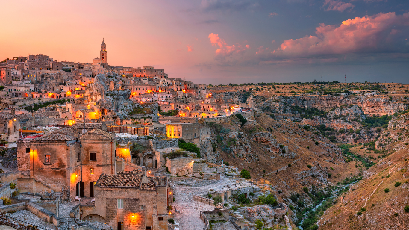 A hillside town at sunset with stone buildings and a tall clock tower, illuminated by streetlights, overlooking a deep valley with rugged terrain and sparse vegetation.
