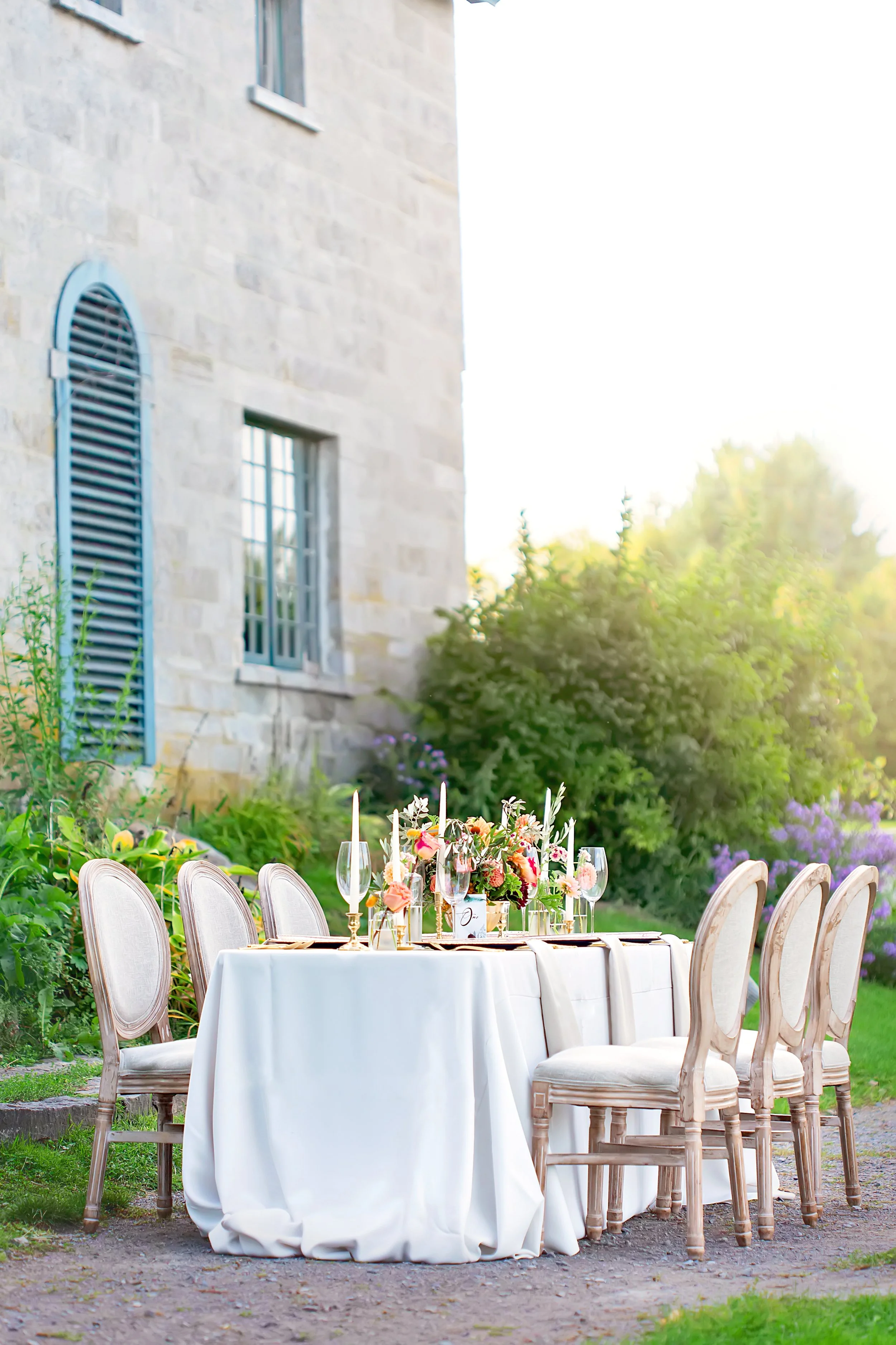 Elegant outdoor dining table with white tablecloth, floral centerpiece, candles, wine glasses, and gold flatware, set in a garden with lush greenery and a stone building in the background.
