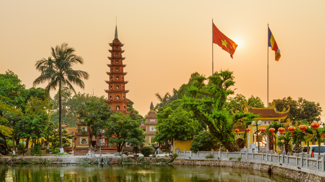 Traditional Vietnamese temple with a pagoda, flags, red lanterns, and a pond at sunset.
