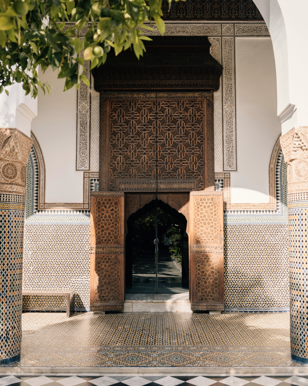Decorative wooden doors with intricate geometric carvings, framed by white walls with ornate tilework and archways, part of a Moroccan or Islamic architectural style.