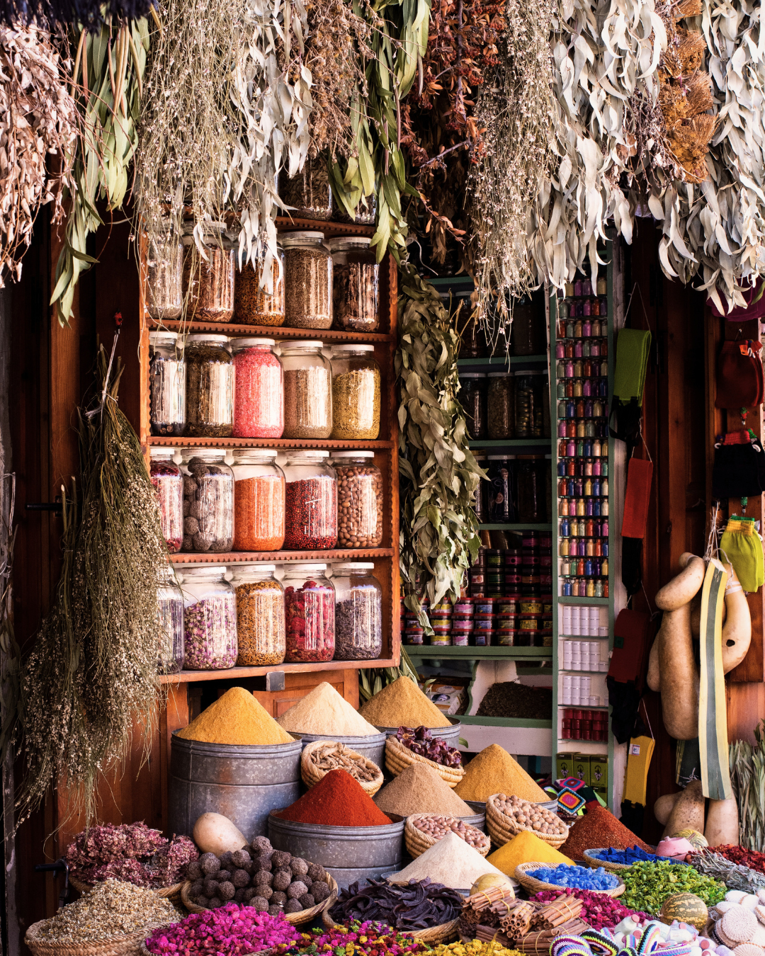 A market stall displays various spices in large metal containers and jars, dried herbs hanging above, and colorful dried flowers and decorative items on the table.