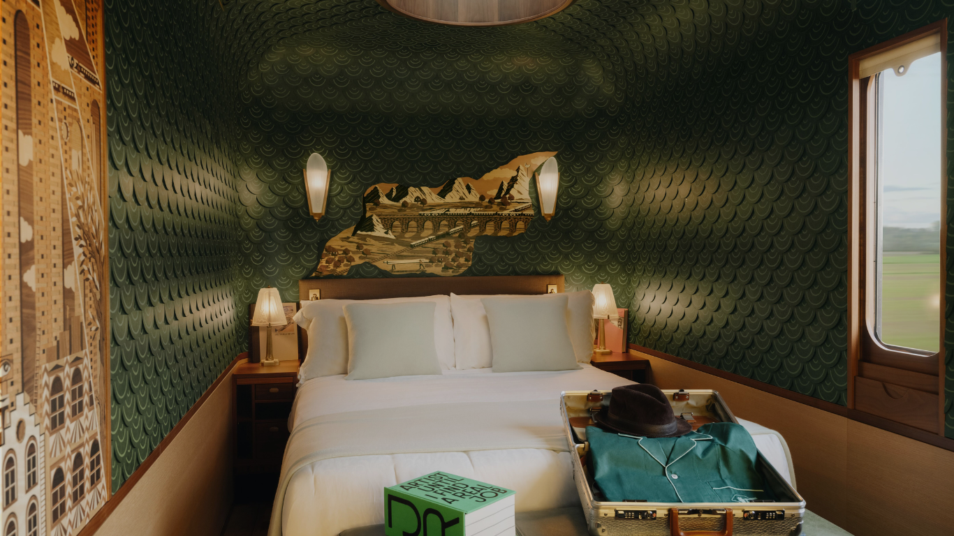 A luxury train cabin with a bed, nightstands, lamps, decorative green wallpaper, and a window showing a rural landscape. There is an open suitcase on the bed containing a hat and clothing.