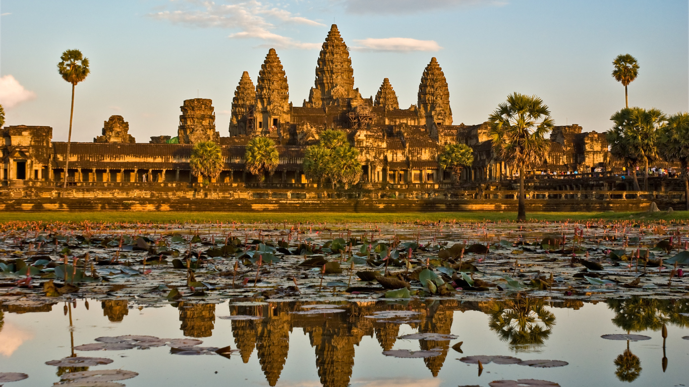 Sunset view of Angkor Wat temple in Cambodia reflected in a pond with water lilies and palm trees in the foreground.