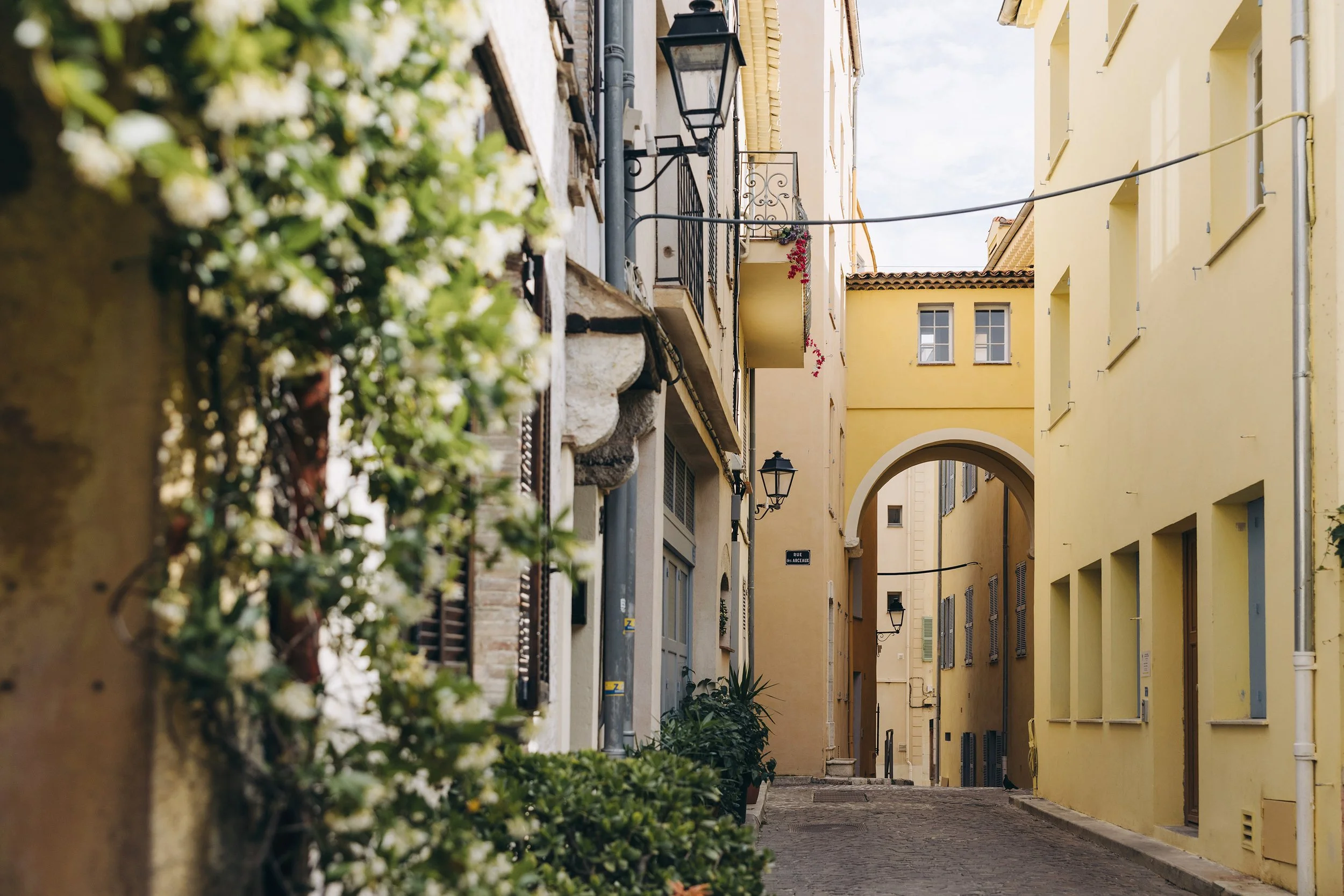 A narrow European street lined with yellow and beige buildings, with balconies, street lamps, and a cobblestone pathway, viewed through some greenery.
