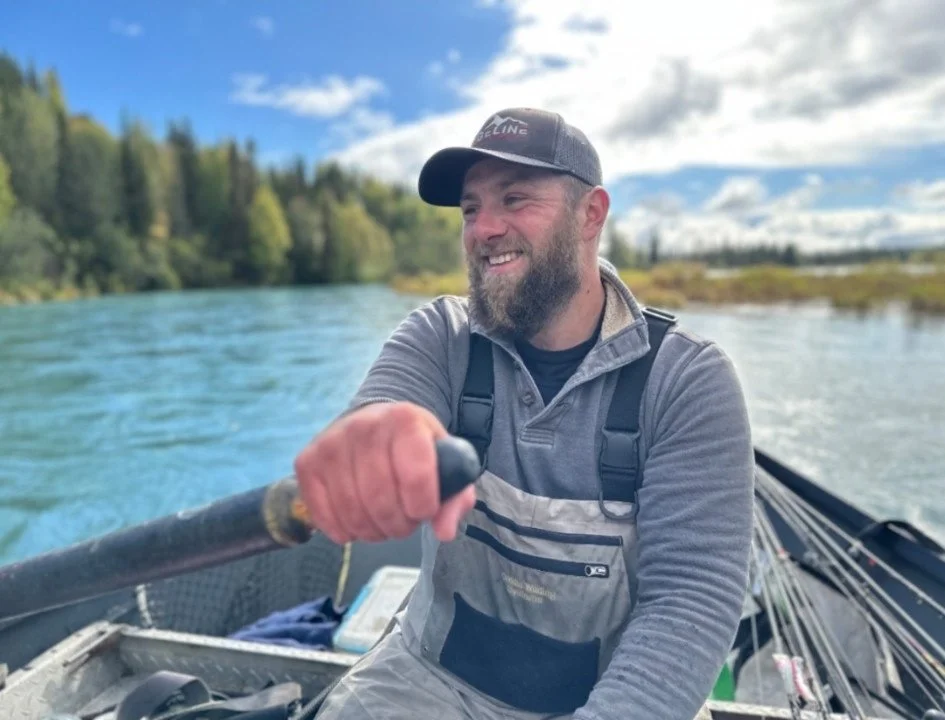 A smiling man in waders holds an oar in a drift boat on a blue-green river.