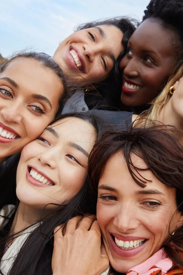 Close-up of six women smiling and hugging outdoors on a cloudy day.