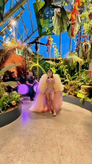 A woman dressed in a pink, fluffy, dramatic gown walking down a pathway in a lush, tropical-themed indoor setting with large green plants and colorful flowers.