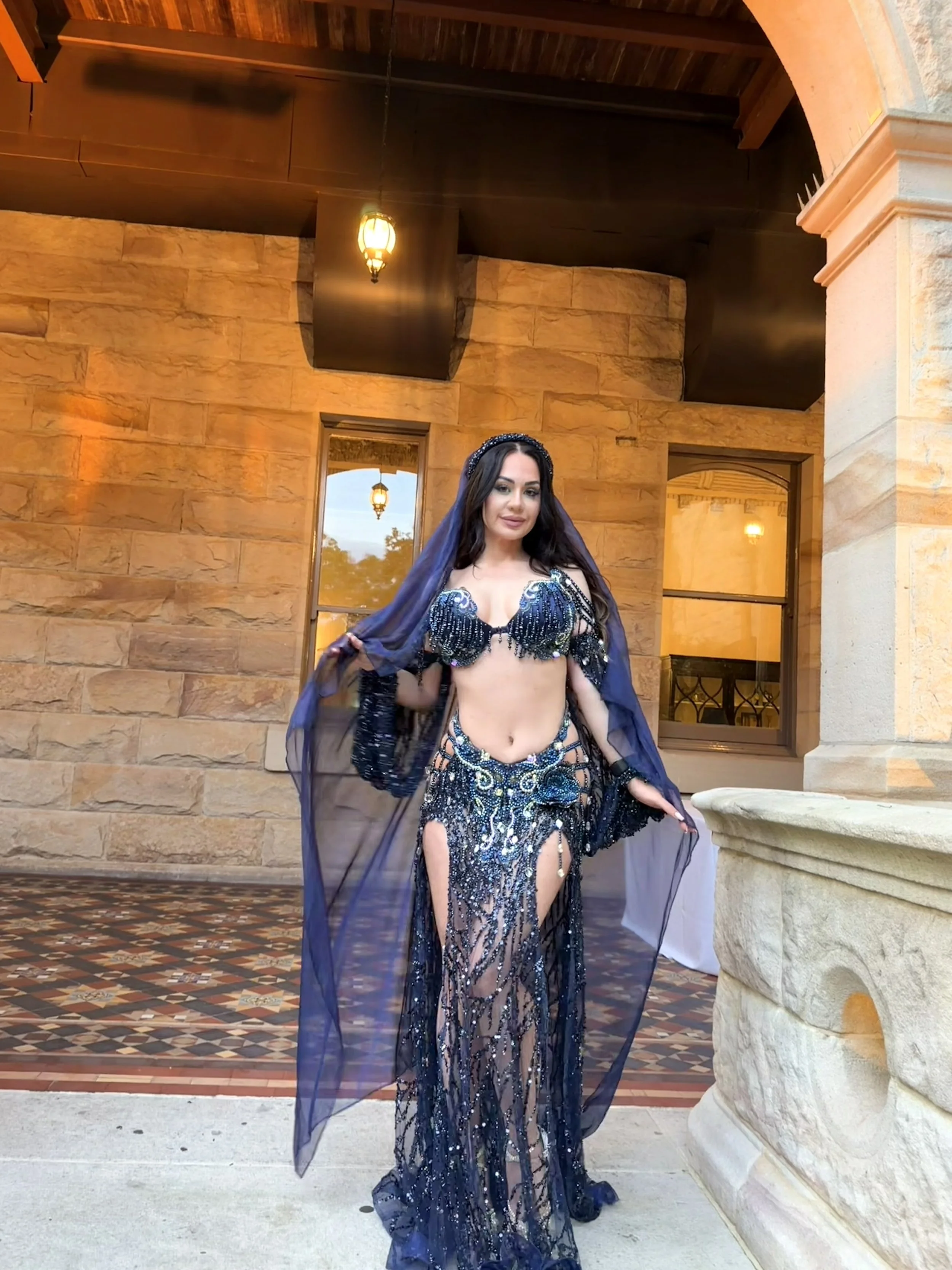 A woman wearing a dark blue, beaded belly dance costume with a sheer skirt, standing outdoors near a stone building with windows, decorative lanterns, and patterned tile flooring, holding the sheer fabric of her costume.