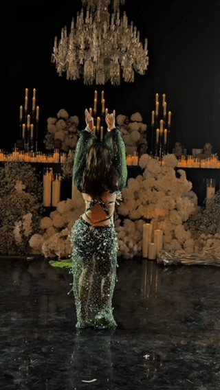 A person performing a dance or pose in front of a decorated backdrop with flowers, candles, and a chandelier.