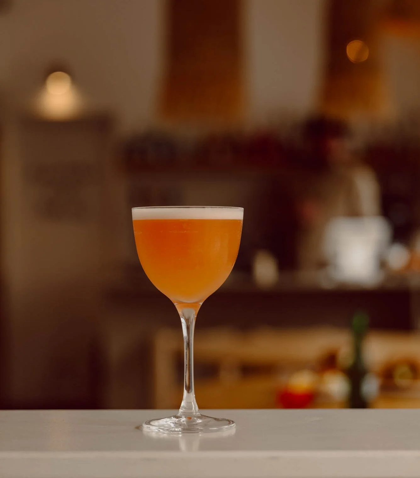 A cocktail glass with an orange-colored drink and a foamy top, set on a white surface in a softly lit indoor setting.