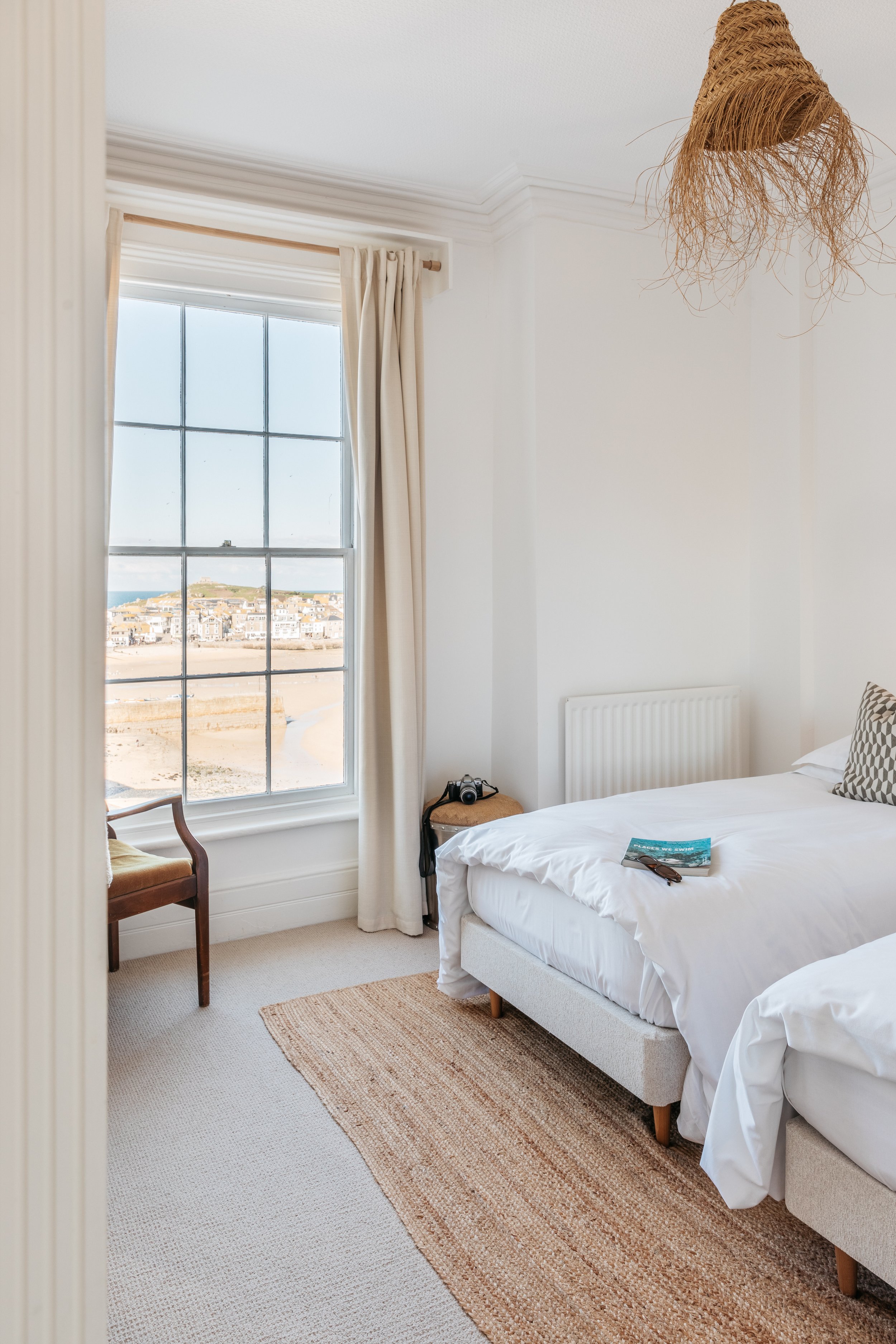 A cozy bedroom with white walls and bedding, a large window with cream curtains showing an outdoor view, a wooden chair, a small side table with a camera, a placed magazine and sunglasses, a woven ceiling lamp, and a beige rug.