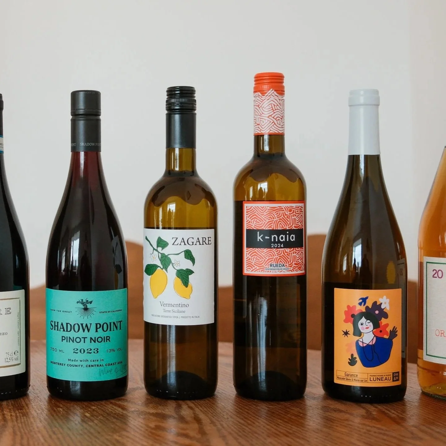A row of 5 wine bottles on a wooden surface, with labels showing different wine types and brands, including Shadow Point Pinot Noir, Zagara Vermentino, k-naia, and others.
