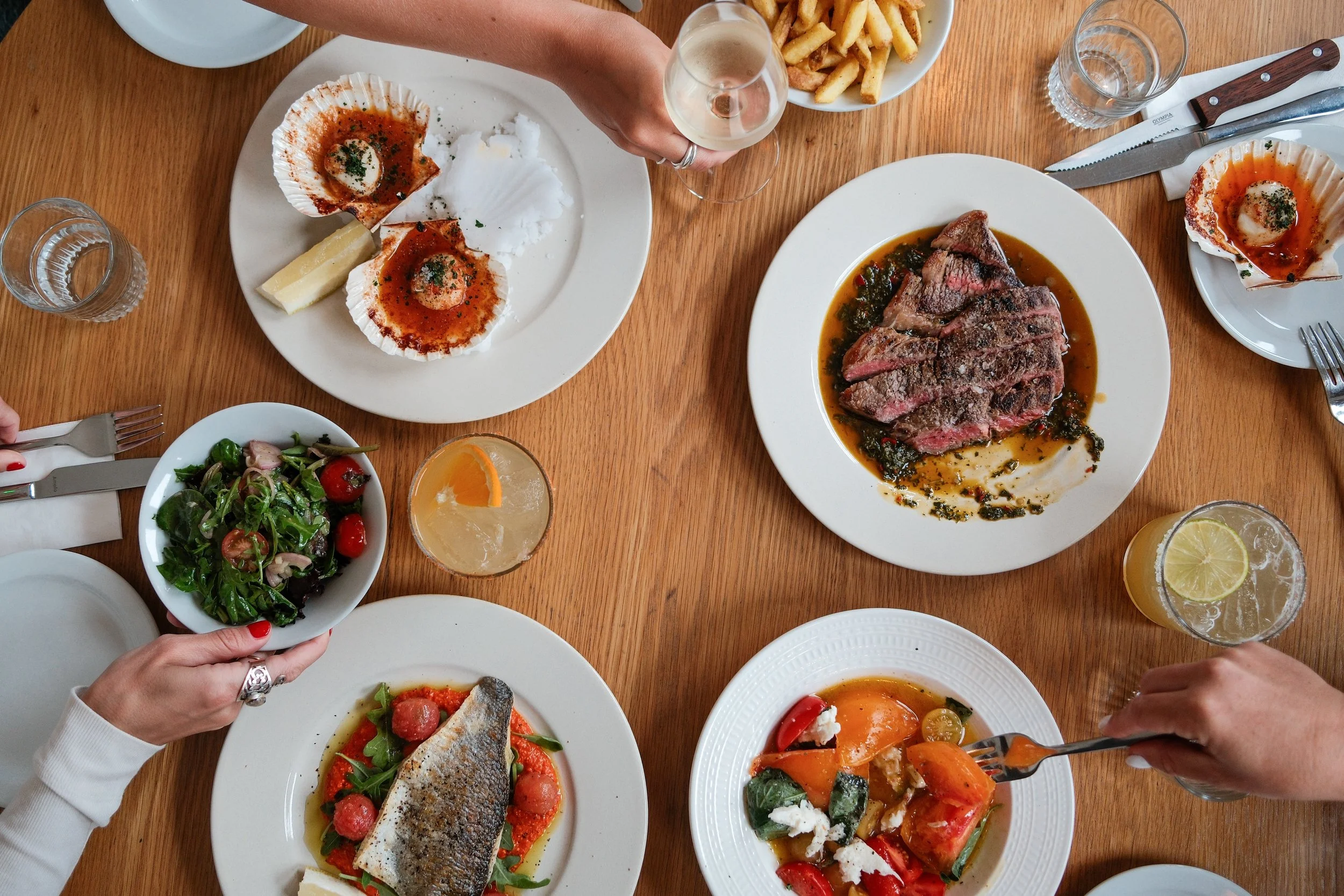 A variety of dishes on a wooden table, including a salad, grilled fish, steak, and side dishes, with glasses of drinks and utensils.