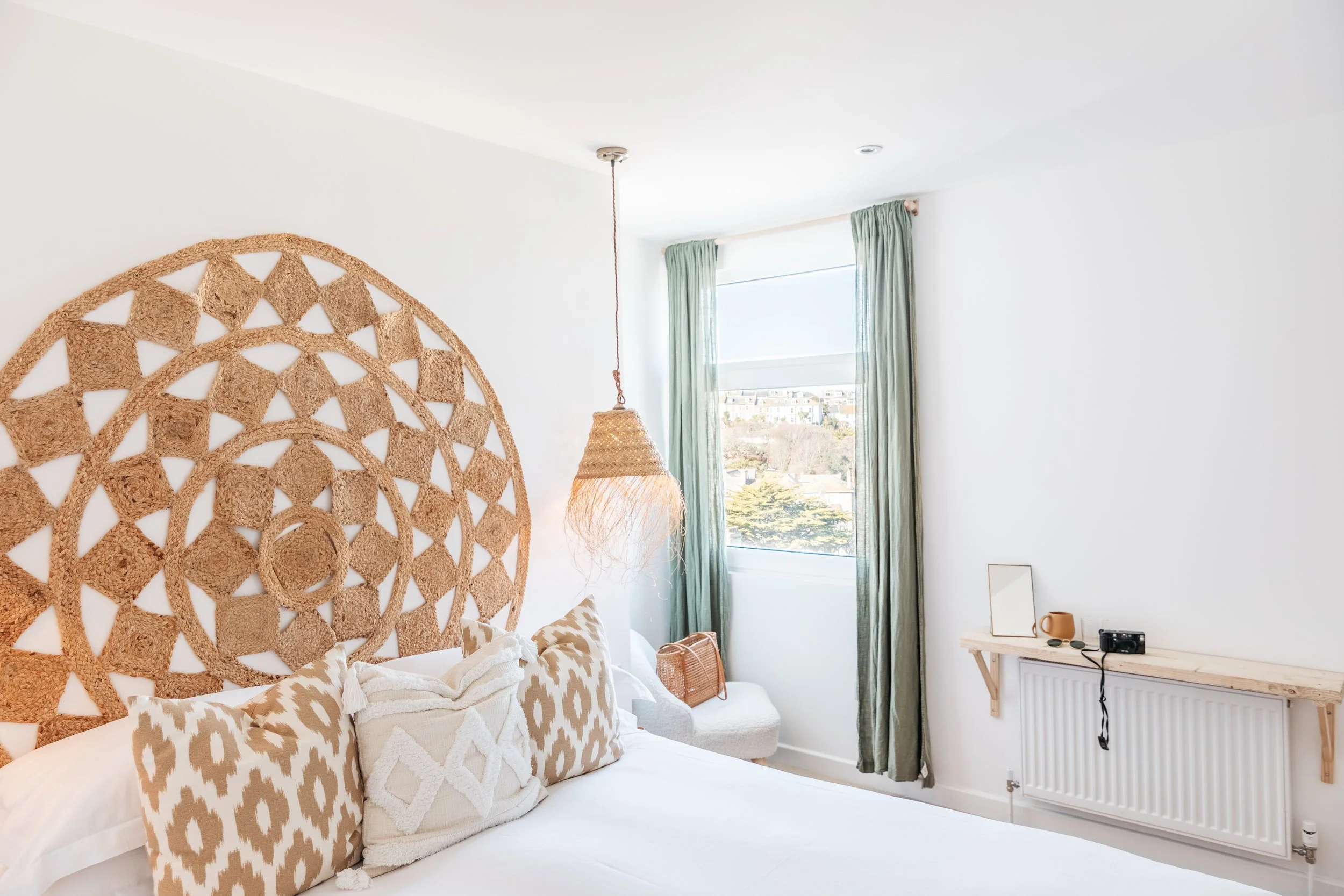 Bright bedroom with a woven headboard, white pillows, and green curtains by a window.