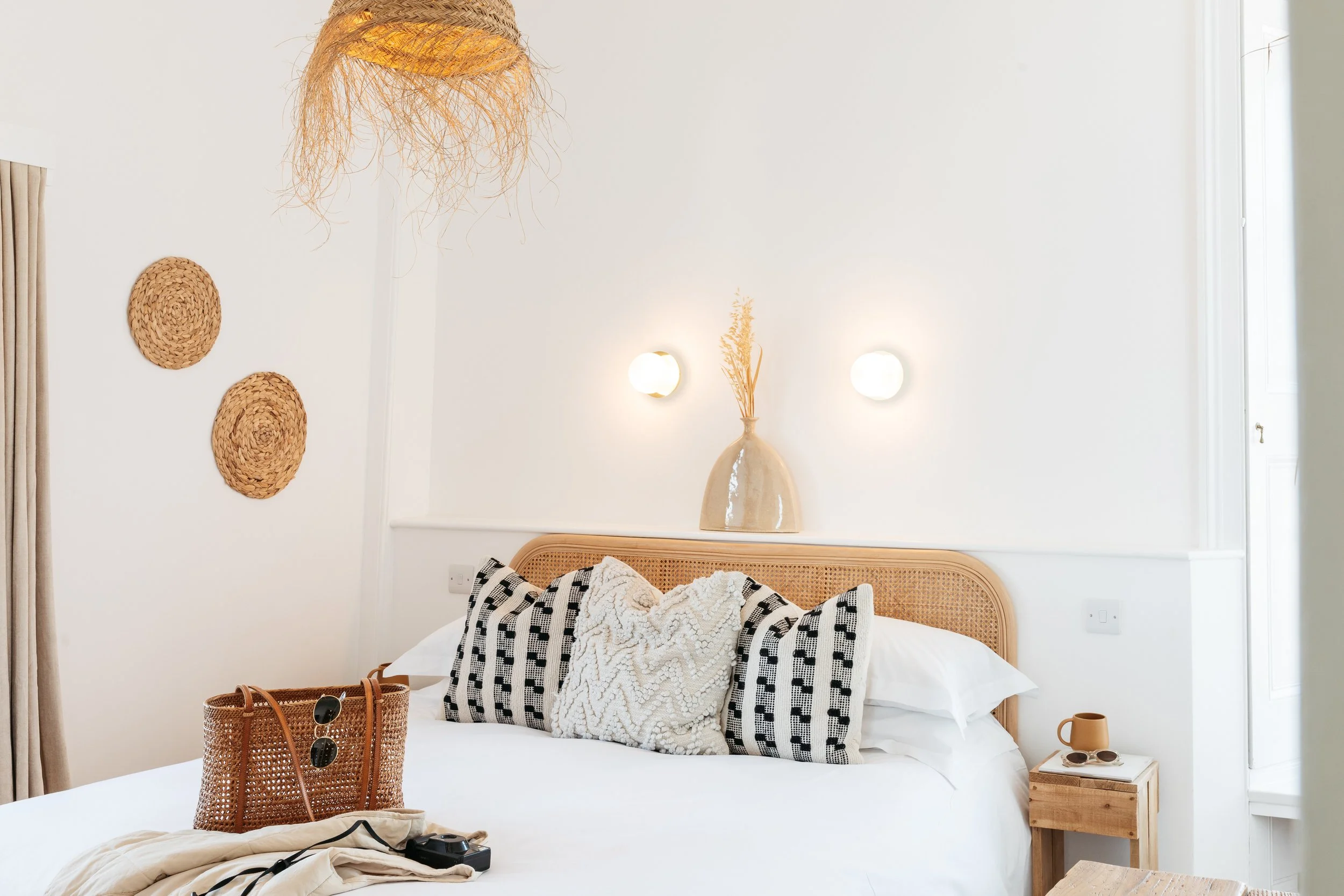 A cozy, well-decorated bedroom with a rattan headboard, white bedding, and decorative pillows, with wicker wall art and a hanging wicker light fixture.