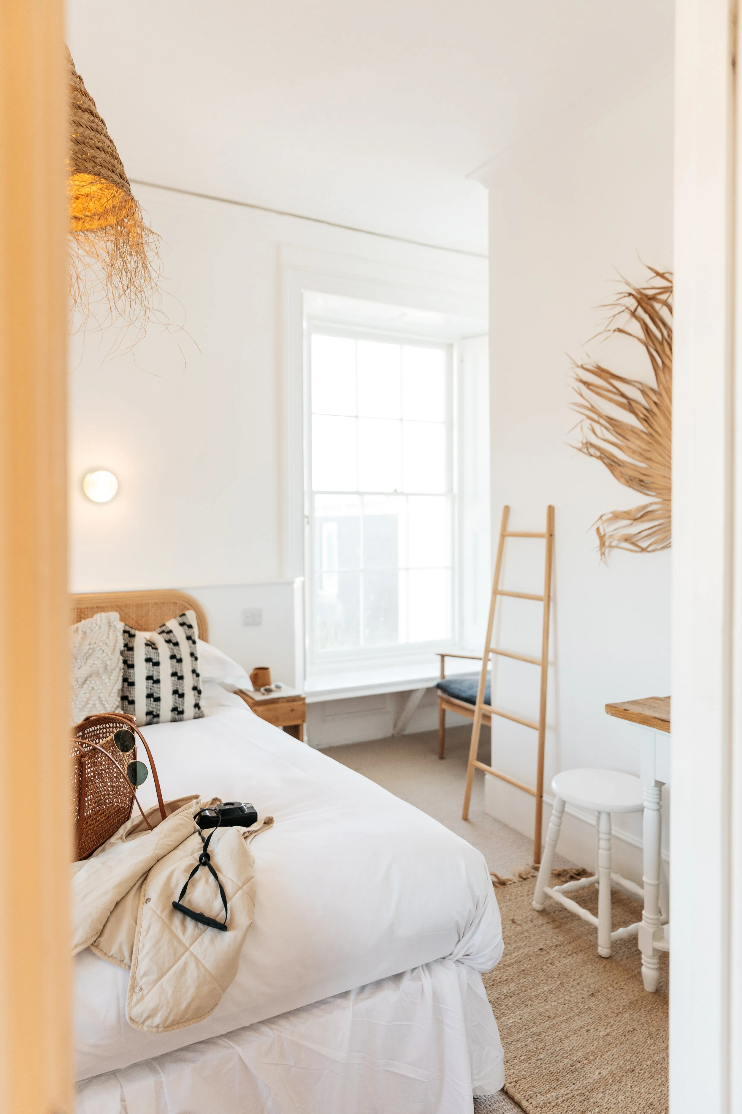Cozy bedroom with white walls and a large window, featuring a rattan bed with white bedding, decorative pillows, a nightstand, a wooden ladder, a small chair, and beach-themed decor.