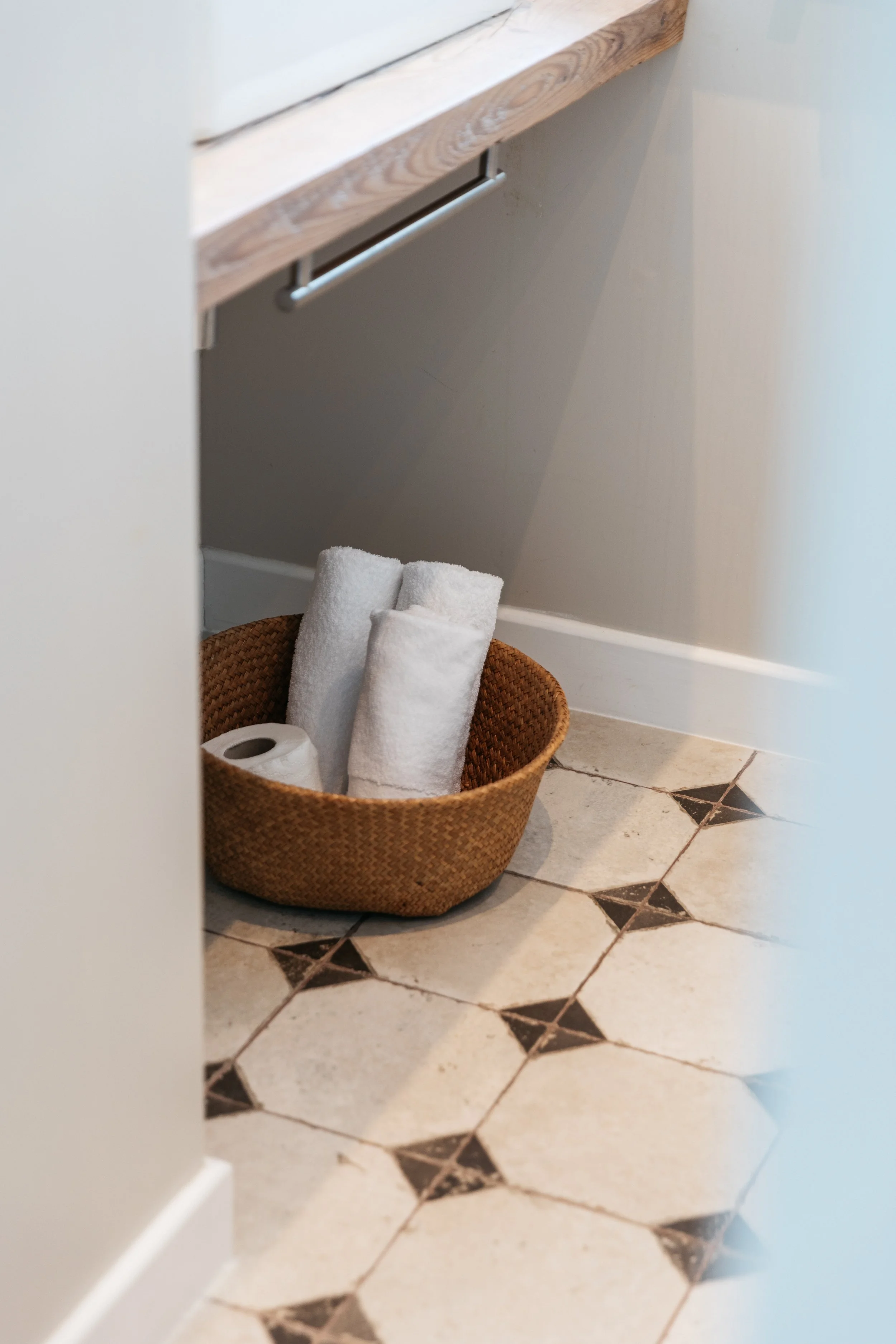 A woven basket containing rolled white towels and a toilet paper roll, placed on a tiled bathroom floor near a cabinet with a wooden countertop.