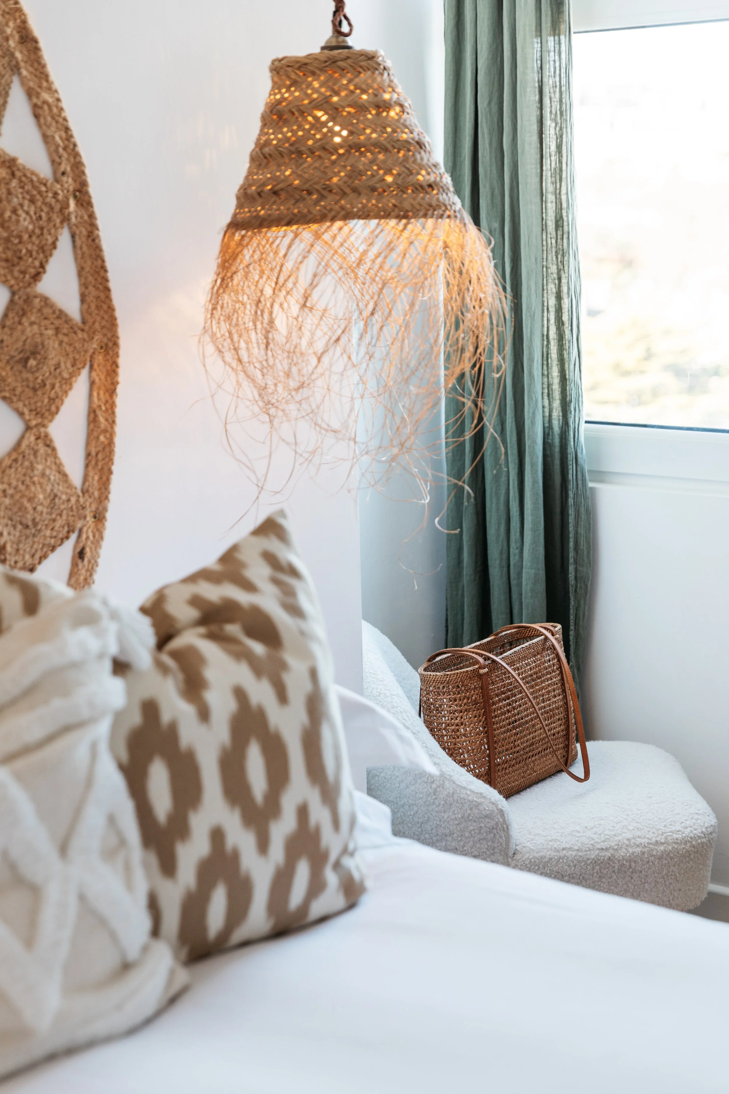 Decorative bedroom corner with a woven hanging lamp, patterned throw pillows, a wicker handbag, and a window with green curtains.