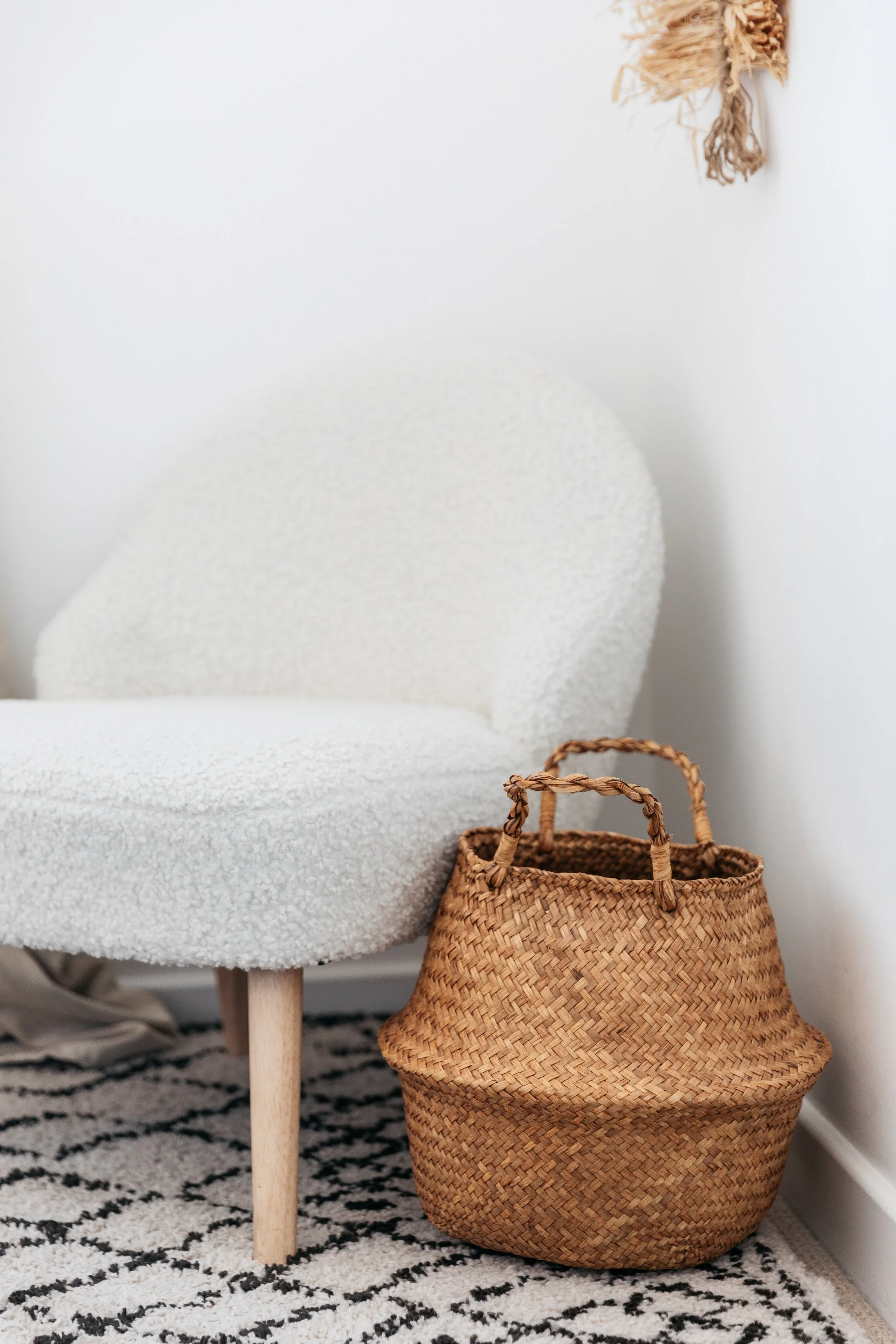 White textured armchair next to a woven straw basket with two handles on a black-and-white patterned rug against a white wall with a hanging woven decoration.