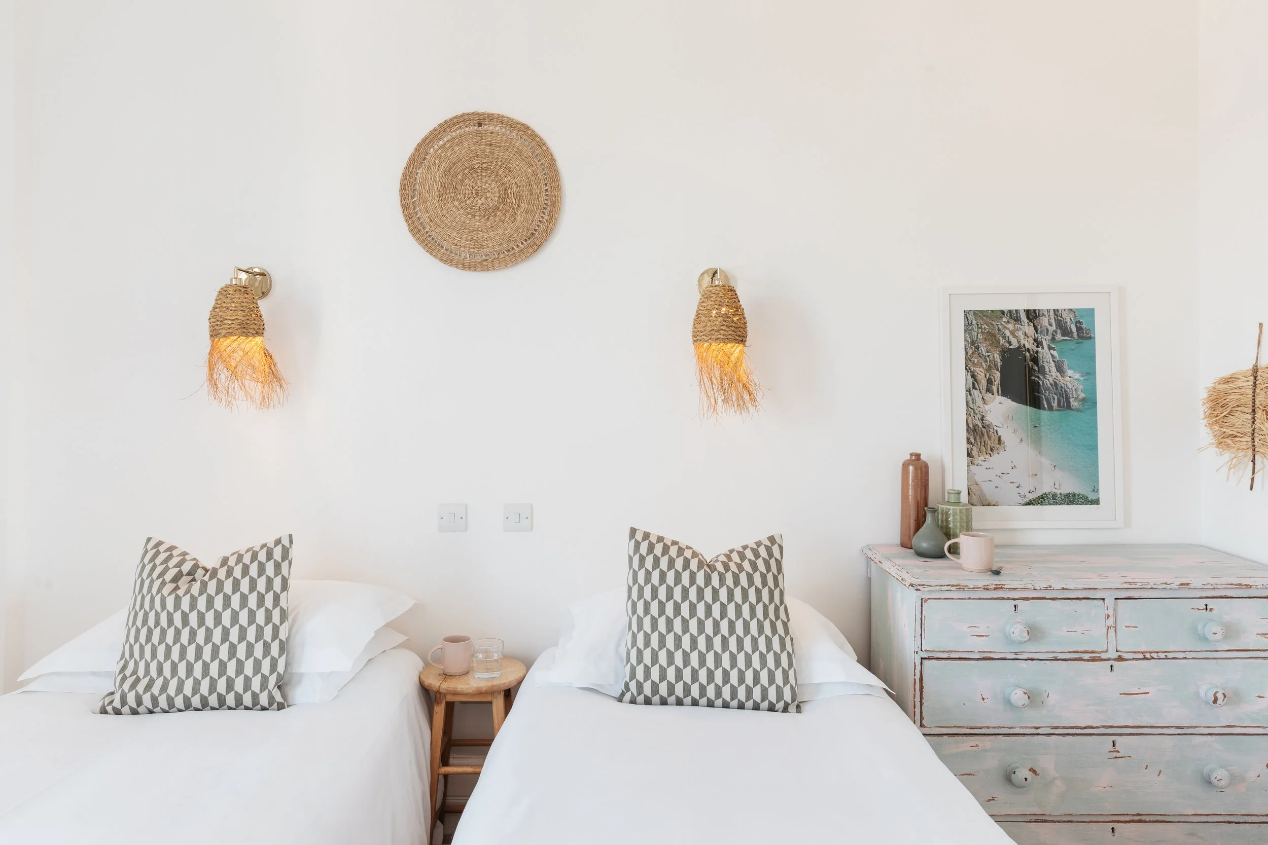 A cozy bedroom with two twin beds, each with patterned pillows, a small wooden nightstand with a mug and glass, wall-mounted woven lamps, a distressed white dresser with vases and a framed beach photograph, and woven decor on the wall.