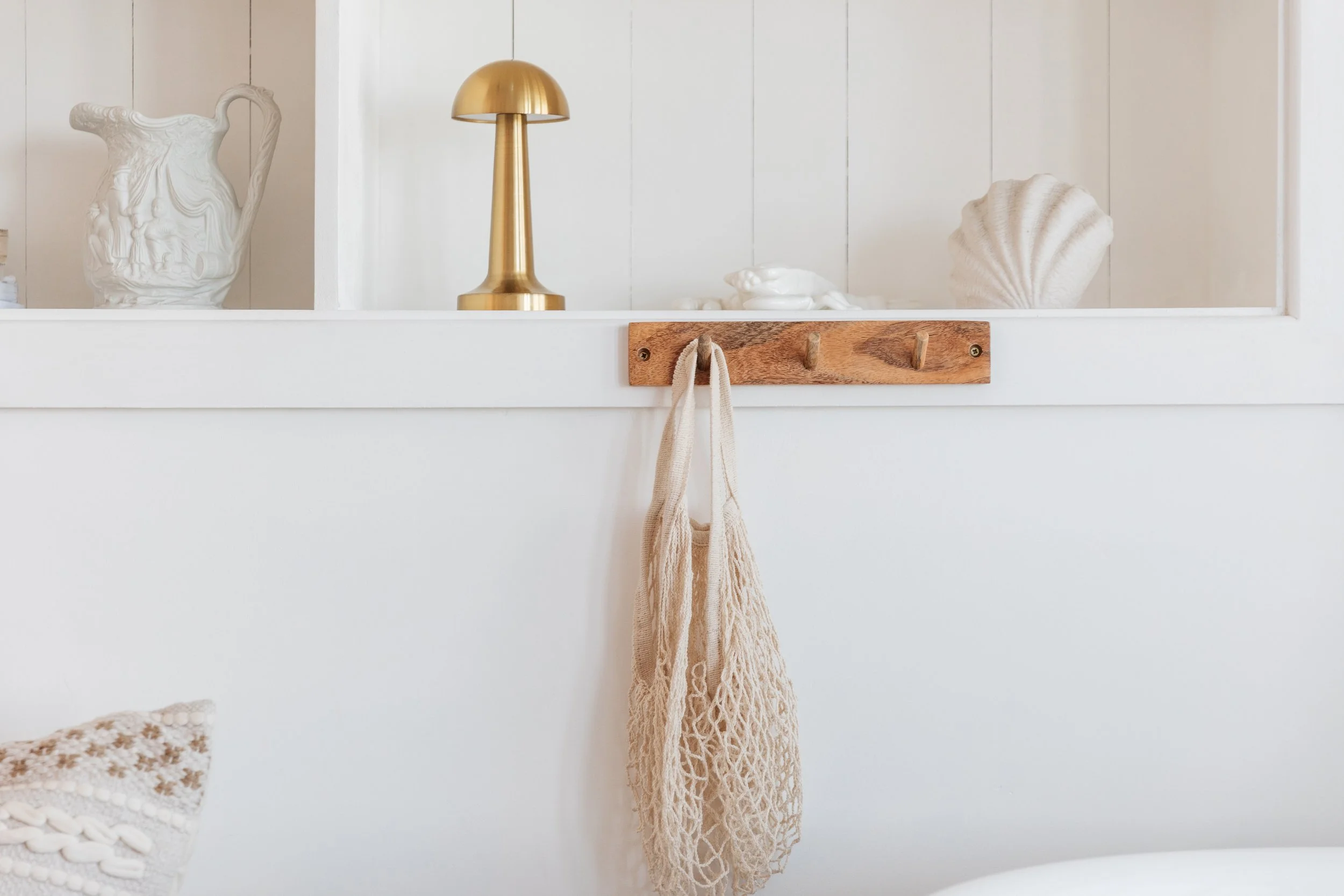 Decorative shelf with white ceramic pitcher, gold table lamp, seashells, and coral, with a beige tote bag hanging from hooks on a wooden board, against white wood-paneled wall